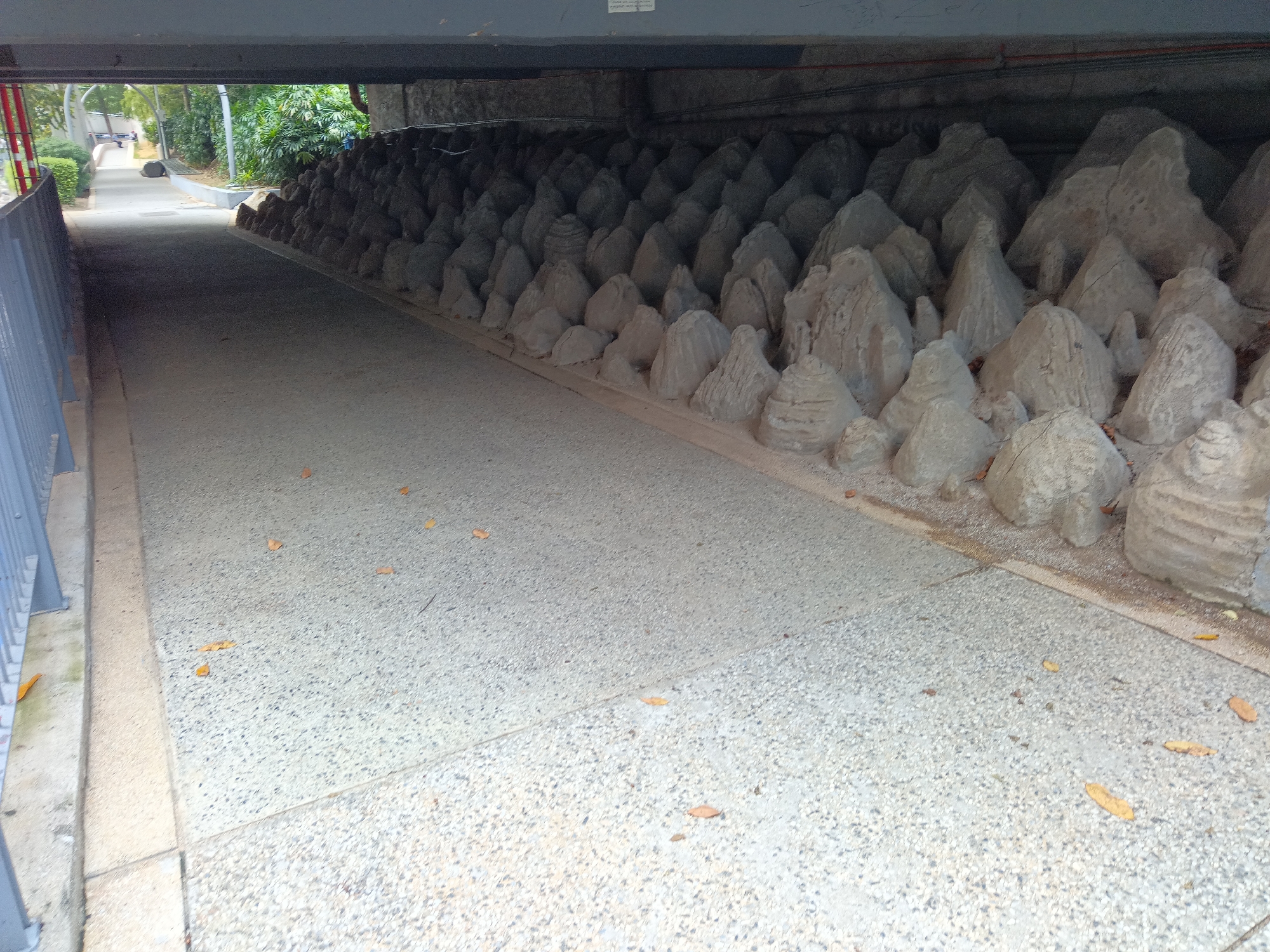 The image shows a pathway under an overhead structure, likely a bridge or underpass, in Kuala Lumpur, Malaysia. The pathway is made of a speckled, gray concrete material and is lined on the right by a dense arrangement of small, conical, gray rock-like sculptures. These sculptures are placed closely together, forming a textured wall. On the left, a gray metal railing runs along the edge of the pathway, separating it from a patch of greenery and another, narrower concrete path that recedes into the distance. The overhead structure is a dark gray, concrete beam. The scene is devoid of people. The lighting suggests it is daytime, with shadows cast by the overhead structure. Scattered on the pathway are a few dry, fallen leaves.