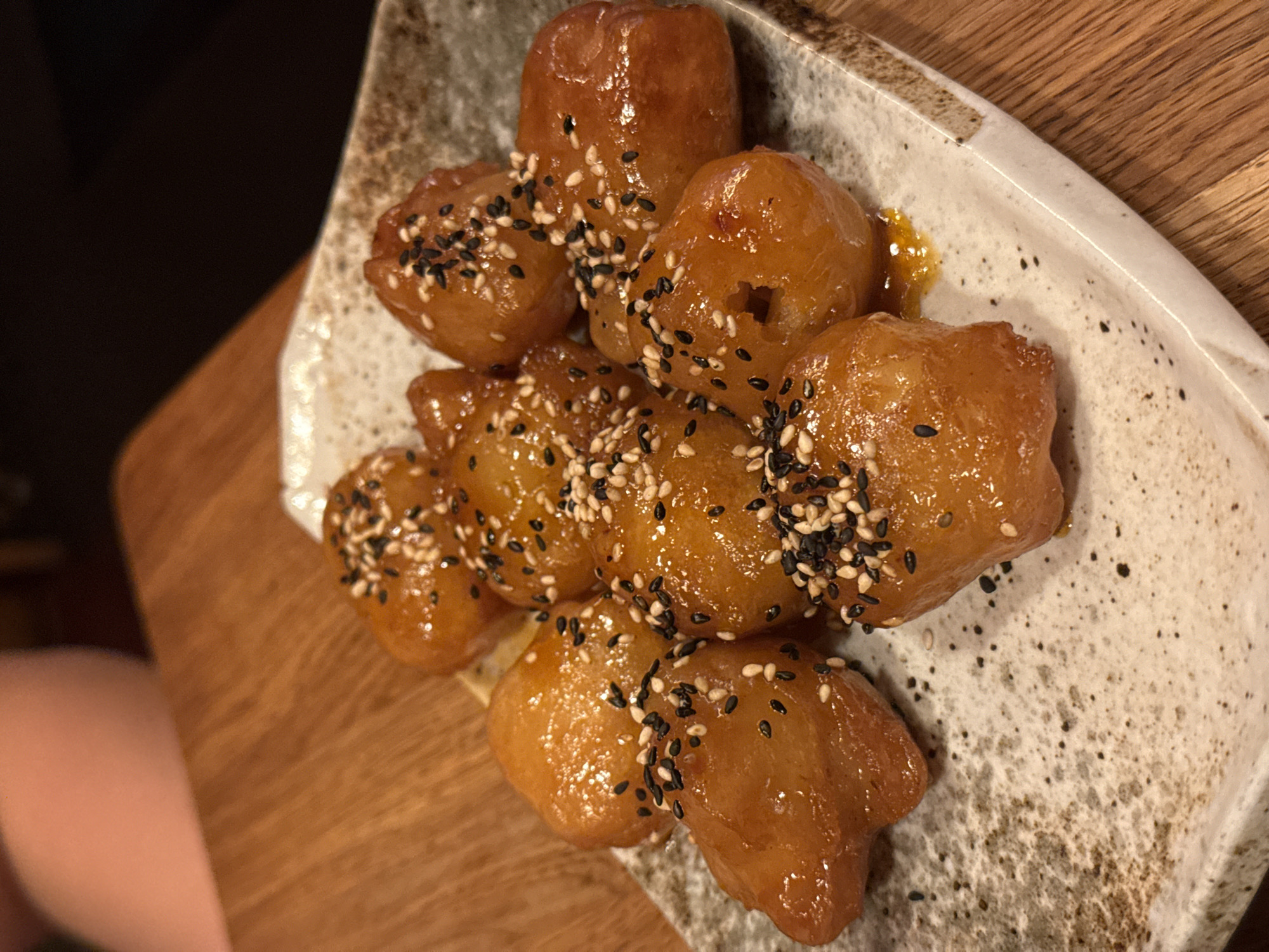 A high-angle, close-up shot depicts a serving of loukoumades, traditional Greek doughnuts, on a speckled ceramic plate. The loukoumades are golden brown and shiny, suggesting they are glazed or syrup-covered. They are sprinkled with black and white sesame seeds. The plate rests on a wooden surface. The scene appears to be indoors. The location information (Sofia, Bulgaria) is not discernable from the visual content of the image.