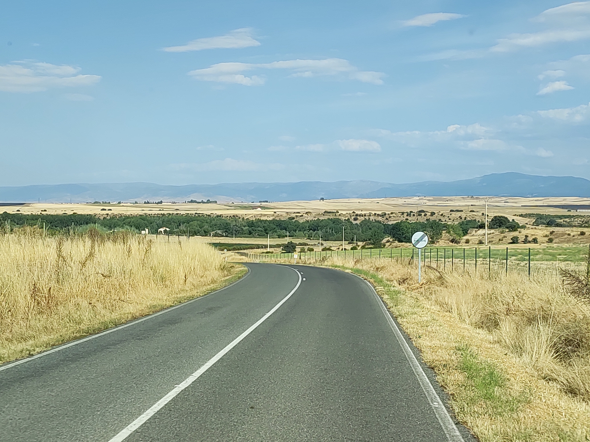 The image shows a paved road curving to the right, lined with dry, golden grass. In the distance, a vast landscape unfolds with cultivated fields, scattered trees, and rolling hills under a bright blue sky with scattered clouds. A fence runs along the right side of the road, separating it from a grassy field. A circular traffic sign with a red border and a diagonal line through it, indicating the end of a restriction, is visible next to the fence. The scene appears to be in a rural area, possibly on the outskirts of Lastras del Pozo, Spain. There are no people or animals visible in the immediate vicinity of the road. The overall impression is one of a quiet, sunny day in the Spanish countryside.