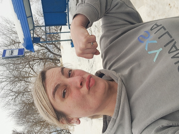 A fair-skinned female is depicted in a close-up, selfie-style photograph taken in the city of Sasovo, Russia. She has blonde hair, pulled back, and her face shows pursed lips and a slightly raised left eyebrow. She wears a grey hooded sweatshirt with the partial, inverted text "SKY ALLIAJ" visible on the chest. Her right arm is bent at the elbow, with her hand fisted and held close to her shoulder in a flexing pose, revealing a ring on her right ring finger. A small silver stud earring is present in her left ear. The background features a blue-framed bus stop shelter with a snow-covered roof, partially visible to the right. Above and slightly behind the shelter, a blue and white rectangular sign with a white bus icon clearly indicates a bus stop. The ground and distant areas are covered in snow, and bare deciduous trees are visible against an overcast sky, indicating a winter environment.