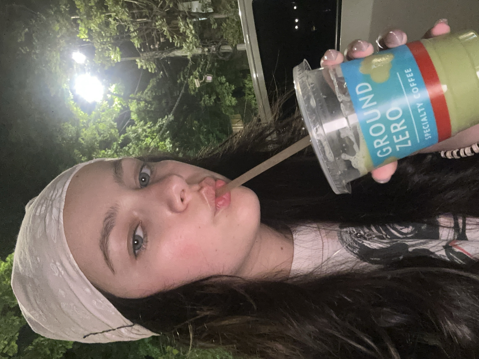 A young woman is outdoors at night, sipping from a large beverage container. The container is labeled "GROUND ZERO SPECIALTY COFFEE" with a green liquid visible inside. She is wearing a white headscarf, a patterned t-shirt, and has dark hair. A straw is in her mouth. The background includes trees, a street light, and a car. Yerevan, Armenia cannot be determined from this image.