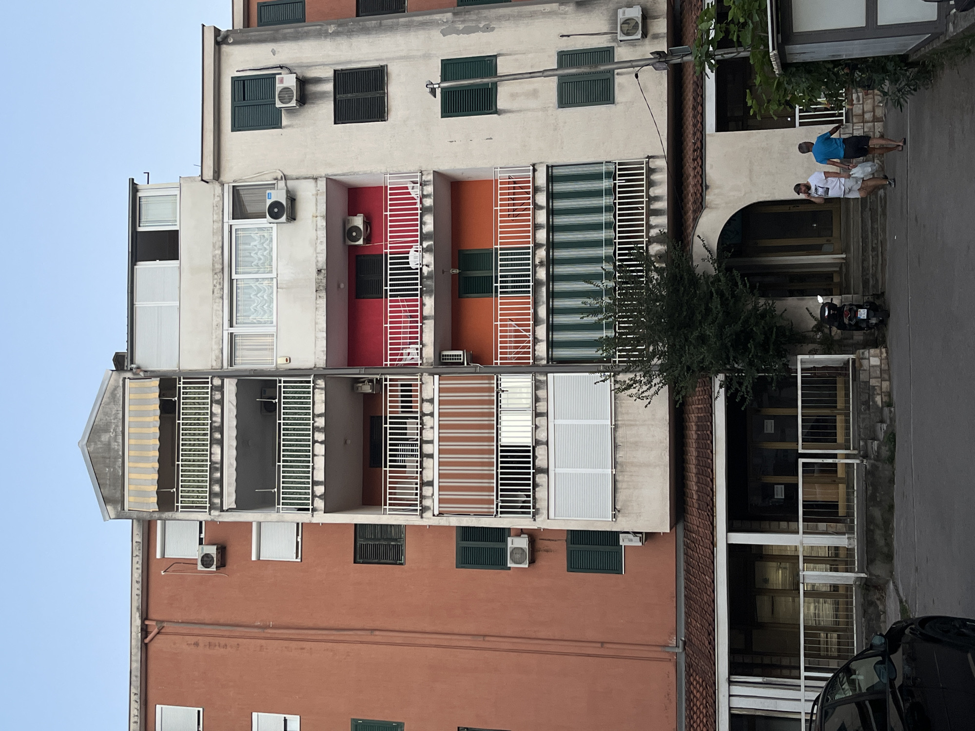 The image shows a multi-story apartment building in Budva, Montenegro. The building has a coral-colored facade on the left and a cream-colored facade on the right, with varying levels of balconies and windows. Several air conditioning units are visible on the exteriors of the apartments. The ground floor of the building features commercial spaces with large glass windows and awnings. In front of the building, a dark-colored car is parked on the left. Two men are walking towards the entrance of the building from the right, carrying shopping bags. A motorcycle is parked to the left of the entrance. The sky is clear and blue.