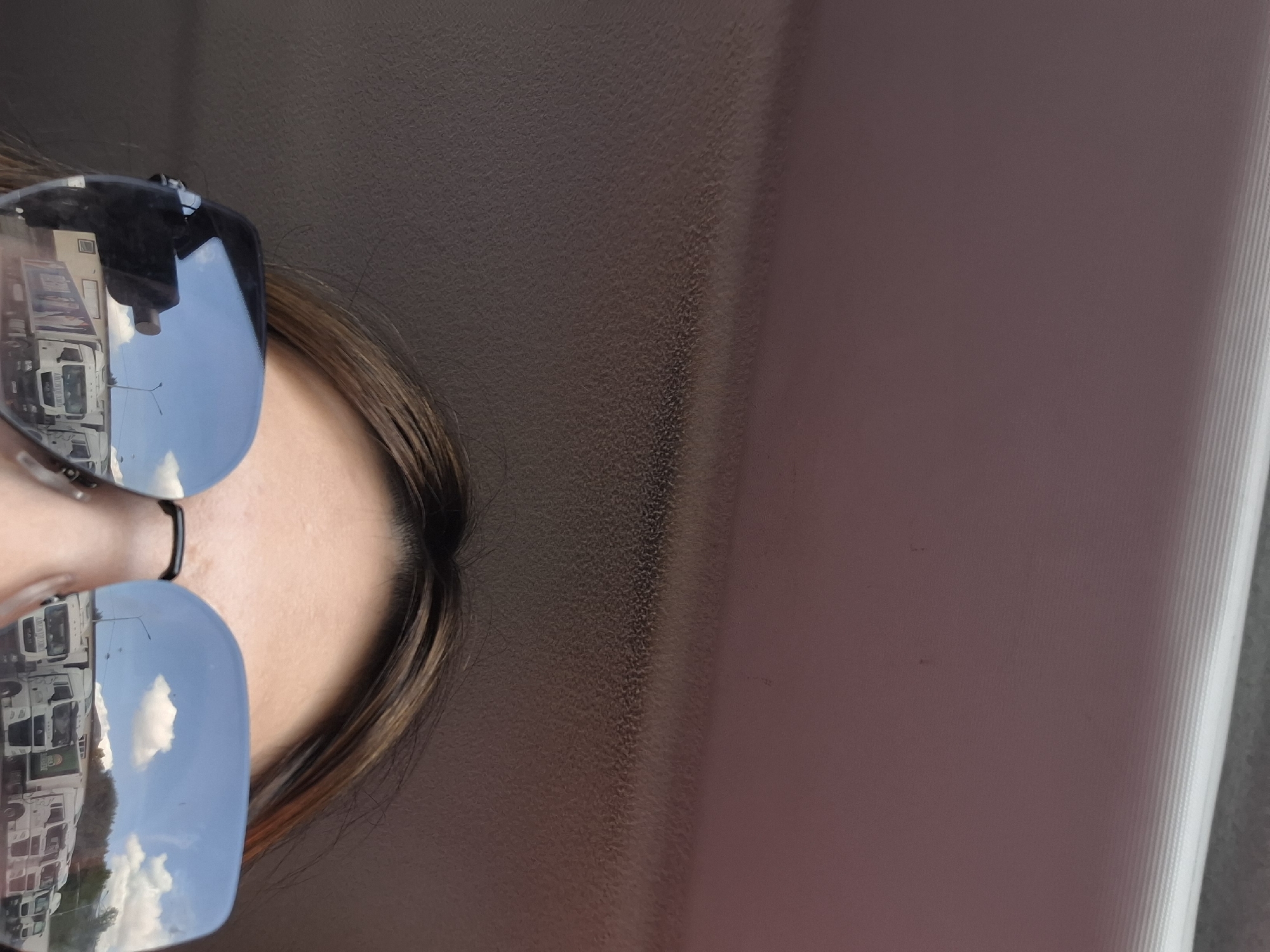 The image presents a close-up, rotated view, primarily featuring the upper part of a person's face obscured by sunglasses. The person has dark brown hair, appearing to be parted in the middle with some lighter brown highlights. Only the forehead and areas around the eyebrows are clearly visible.
The central objects are the large, mirrored sunglasses, which reflect a bright outdoor scene. Both lenses show a clear blue sky with fluffy white clouds overhead. Reflected prominently in the sunglasses are several white commercial trucks, appearing to be parked or traveling on a road or lot. Visible text on one of the truck reflections clearly shows "AUMAN," and another truck might display "FORLAND," indicating specific truck brands. A lamppost or utility pole is also part of the reflection.
The background directly behind the person is a dark brown, textured surface, possibly the interior of a vehicle, such as a headliner or dashboard. To the far right, a lighter, vertically ribbed surface, perhaps a window frame or car pillar, is visible.
The scene takes place during the daytime, under sunny conditions with some cloud cover. The activity depicted is simply the person observing the outdoor environment, as reflected in their sunglasses, likely from inside a vehicle. The location context provided is Izhevsk, Russia. No discernible emotions are visible.