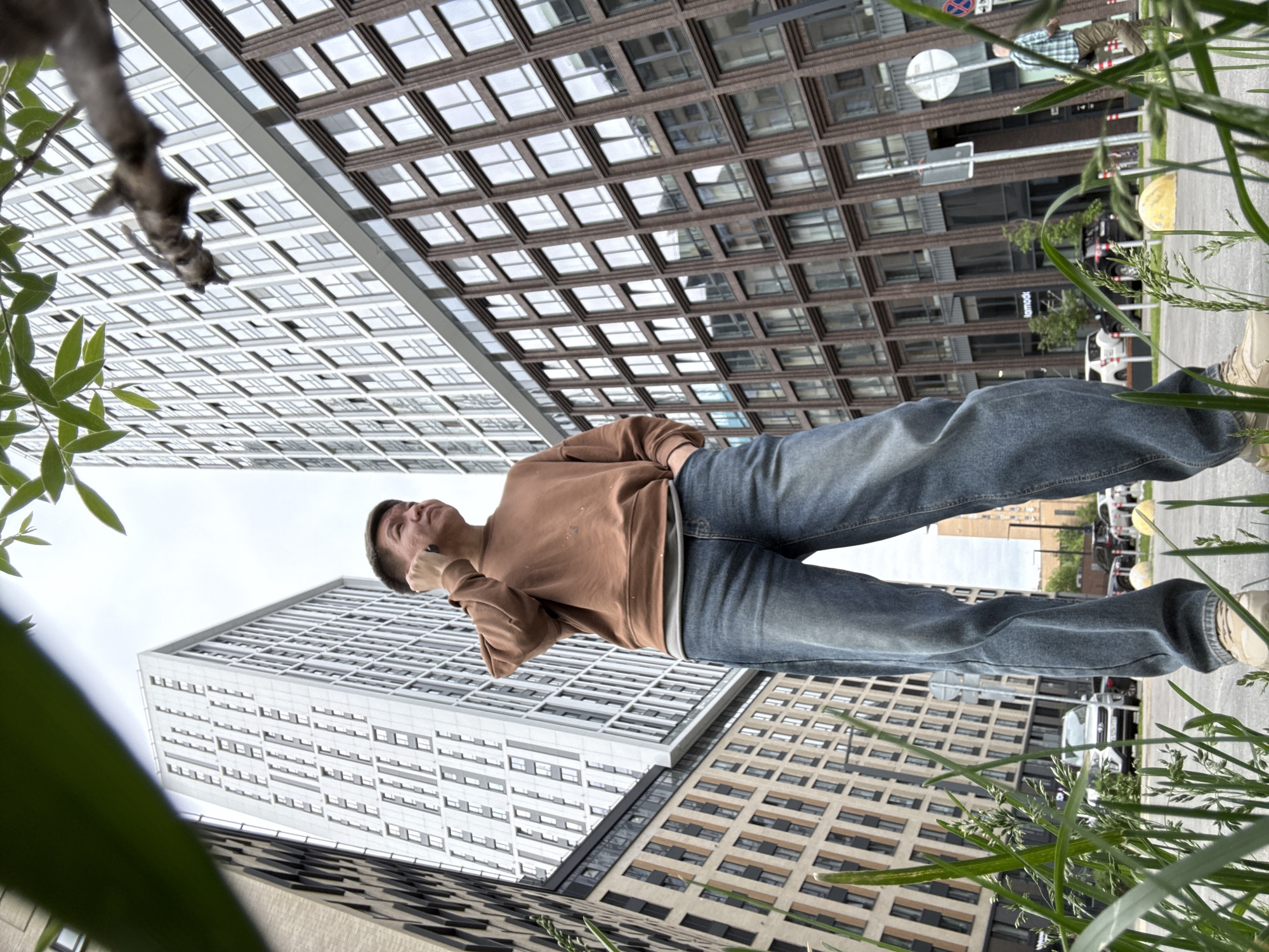 The image captures a low-angle view of a young man looking upwards in an urban setting in Moscow, Russia. The perspective is from the ground, looking up towards several tall, modern buildings under a bright sky.
The central subject is a young man, dressed in a brown long-sleeved sweatshirt, blue jeans, and light-colored sneakers. He stands with his left arm bent, hand near his chin, and is gazing intently upwards, suggesting observation or contemplation of the buildings or the sky. His hair is short and dark.
The background features several large, multi-story urban buildings. To the left, a prominent light-colored building with numerous small, rectangular windows dominates the scene. To the right, another building displays a brownish-grey facade with a distinct grid pattern of square windows. Further back, a beige/brown building with horizontal window strips is visible.
Green foliage, including leaves and blades of grass, partially frames the bottom and left edges of the photograph, adding a natural element to the otherwise concrete landscape. A bare tree branch is also visible in the top left.
In the mid-ground, a street with parked cars can be seen, including a white vehicle. A circular white road sign and a street light pole are also present. The word "HANKOOK" is clearly visible as text on one of the buildings in the background. The lighting suggests it is daytime, with a bright, possibly overcast, sky.