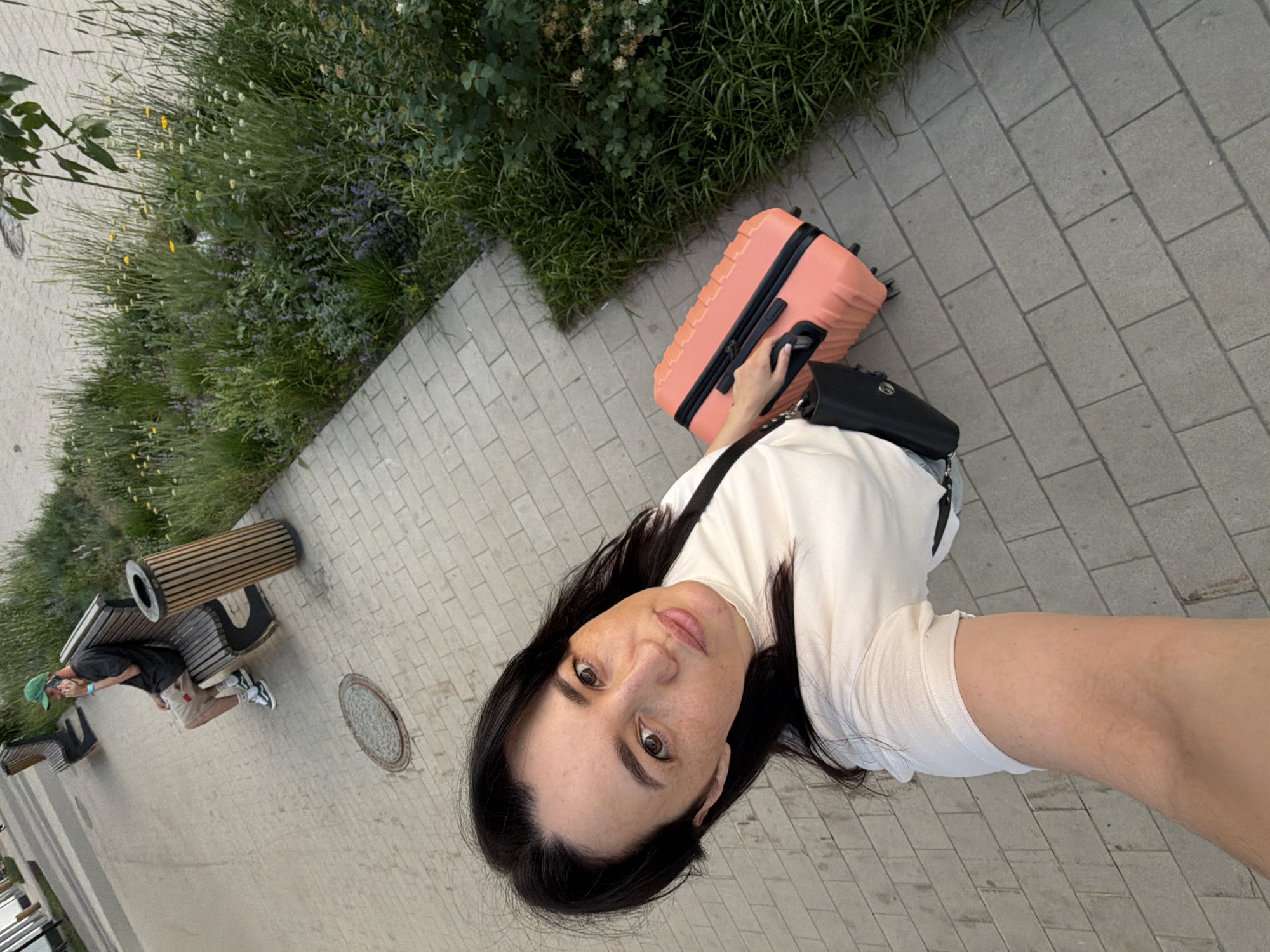 A woman with dark hair and freckles is standing on a paved walkway, holding a pink suitcase and a black purse. She is wearing a white turtleneck shirt and light-colored shorts. In the background, a man in a green cap and beige shorts is sitting on a park bench, talking on his phone. The walkway is made of light gray rectangular pavers. To the right of the walkway is a lush green garden with various plants and flowers. A wooden trash receptacle is also visible near the bench. The scene appears to be in a park or public outdoor space in Moscow, Russia, likely during daytime.