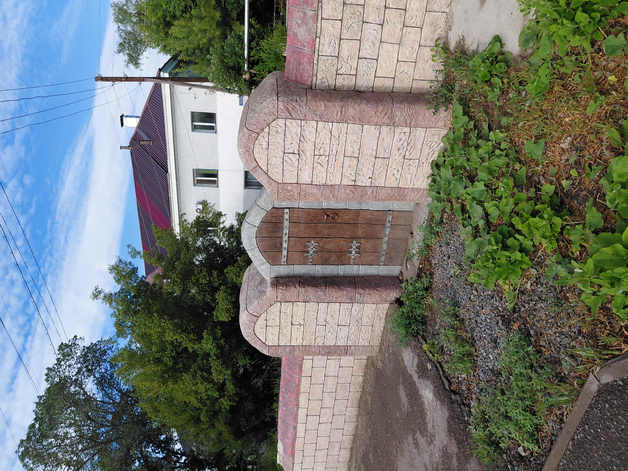 A wooden gate with metal hinges is set into a decorative stone wall in Karaganda, Kazakhstan. The wall is constructed of rectangular stone blocks with a reddish-brown trim along the top. Three rounded pillars with arched tops flank the gate and extend the height of the wall. The gate itself appears to be made of planks of wood with decorative metalwork and a large metal ring handle. Beyond the wall, a building with a red-tiled roof and light-colored walls is visible, along with trees and utility lines against a blue sky with scattered clouds. In the foreground, there is a patch of green plants and gravel next to a paved area.