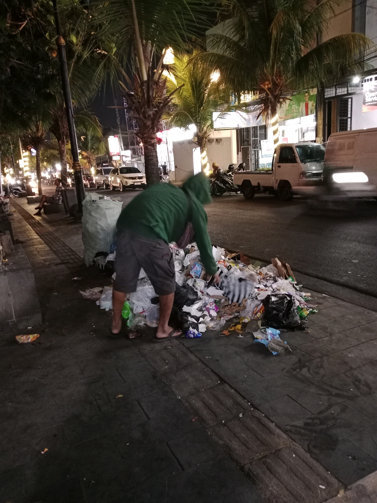 A person wearing a green hooded jacket and gray shorts is bent over a pile of trash on a sidewalk at night. The trash is a mix of plastic bags, paper, and other debris, some of which appears to be spilling onto the street. A large, light-colored trash bag is situated next to the pile. In the background, palm trees line the street, and there are vehicles, including a white pickup truck and a van, as well as buildings and streetlights. The scene depicts a public area with a significant amount of litter. The location is the city of Cianjur, Indonesia.