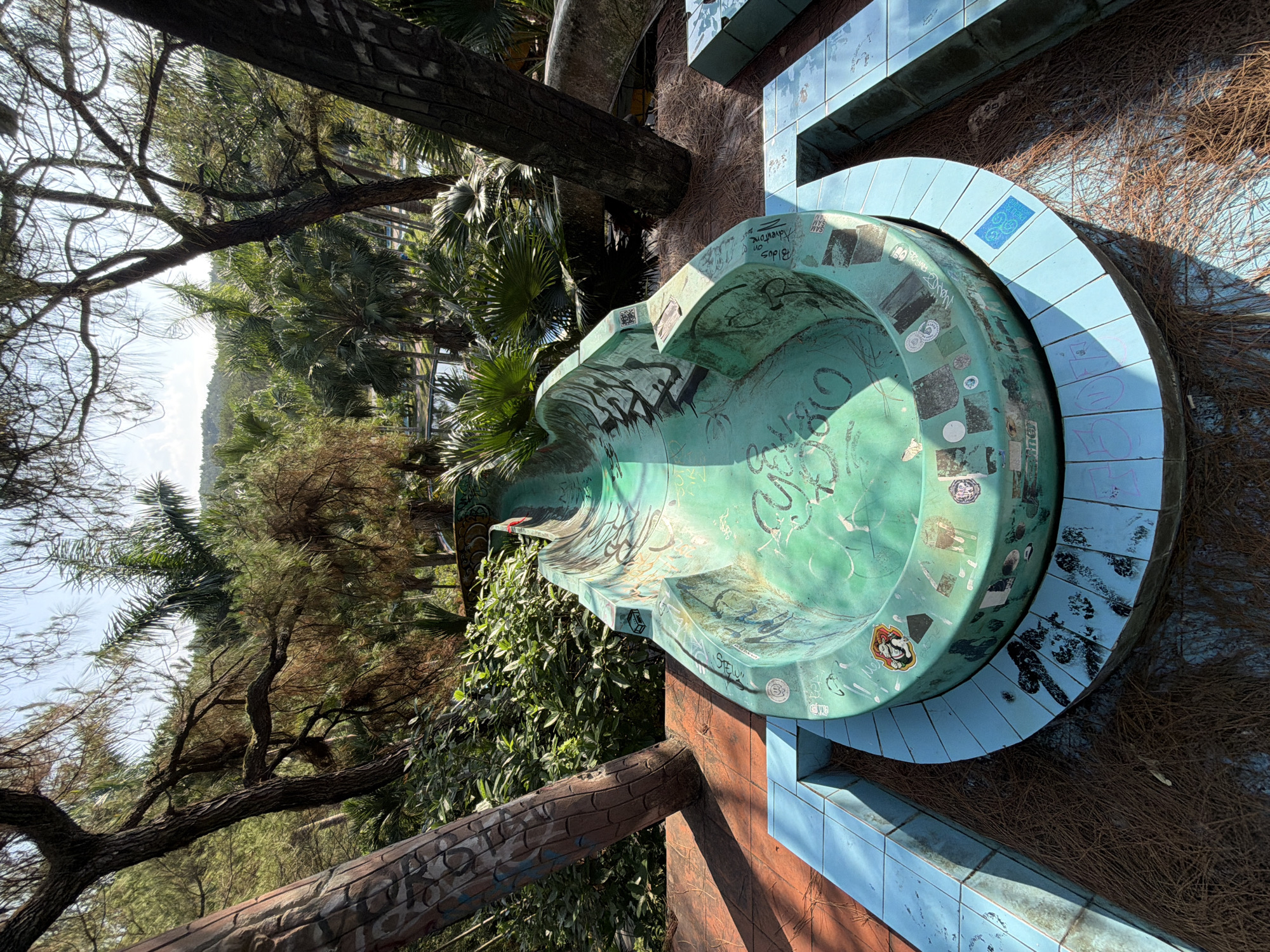 The image shows an outdoor park area in Thủy Xuân Ward, Vietnam. A defunct, turquoise, graffiti-covered fountain is the central object. It is round with a curved, raised lip and surrounded by light blue tile steps. The area is covered in dried pine needles. The background includes dense greenery, primarily trees and foliage. A section of brickwork, possibly part of a wall or other structure, is also visible. The image is taken during daylight hours.