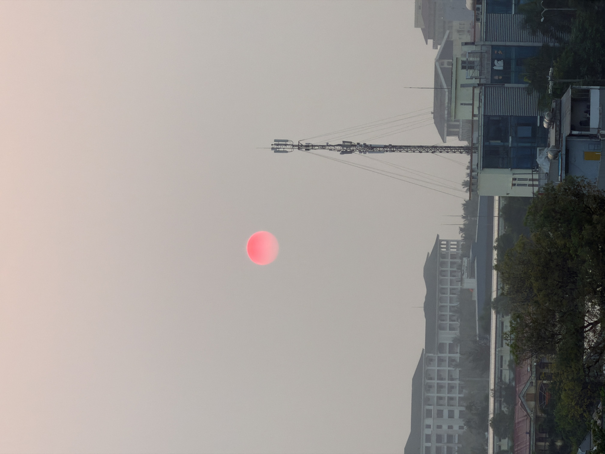 The image shows an urban landscape in Đà Nẵng, Vietnam, likely at dawn or dusk due to the pale orange sky. The sun appears as a reddish-orange disk, low in the sky. A large telecommunications tower dominates the scene, with its lattice structure and multiple antennae. Buildings of varying heights and architectural styles form the skyline, including several mid-rise structures with a grid-like facade. Trees and vegetation are visible in the foreground and background, indicating a mix of urban and natural elements. No people are visible in the image. The overall scene has a hazy or muted quality.