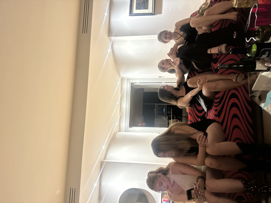 Five women sit close together on a red and black striped sofa. The first woman at the bottom of the image is looking at her phone. The next three are wearing black dresses. The woman at the top is wearing a black dress with jewelry. Wine bottles and other items rest on the sofa next to them. A doorway is visible in the background with a kitchen area beyond. A framed picture hangs on the wall.