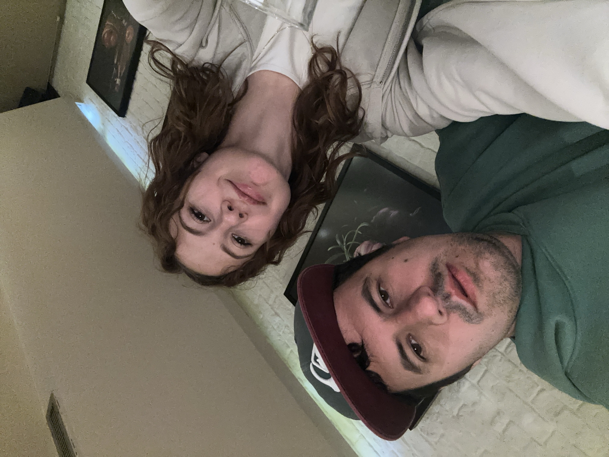 A selfie taken from an unusual angle shows a man and woman indoors. The woman has red, curly hair and is wearing a white hoodie. The man is wearing a green hooded sweatshirt and a baseball cap with a maroon bill. Two framed pictures are visible on the wall behind them. The location is unknown.
