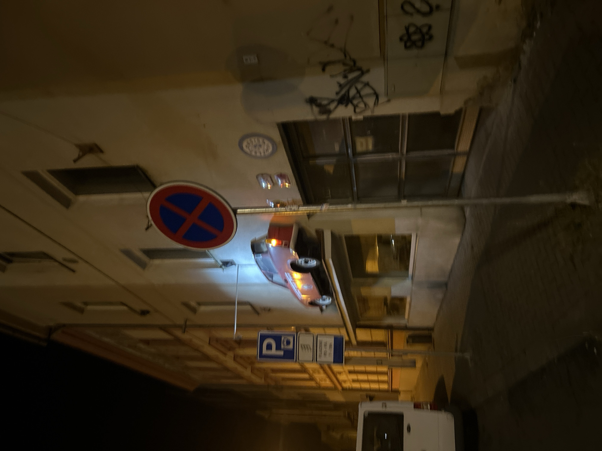 The image shows a street scene at night in Pilsen, Czechia. A vintage pink car is mounted on the exterior wall of a building. It is positioned above a commercial window. A blue and red "no parking" sign is visible near the car. Below the car, a blue parking sign with a 'P' is also present. A white van is parked on the left side of the image. The building itself is light-colored with multiple windows. Graffiti is visible on the lower section of the building. The street appears to be paved with cobblestones.