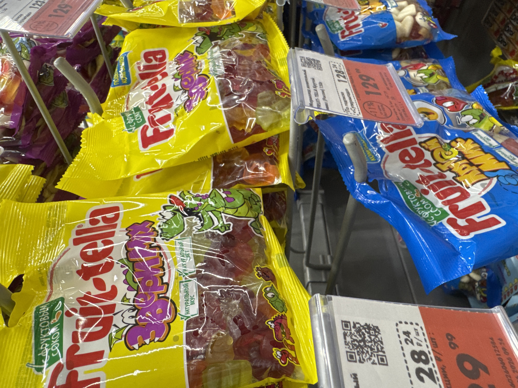 This is a close-up, eye-level shot of a grocery store display featuring packages of gummy candies. The focus is on two main types of packaging: bright yellow bags with a large, colorful "Frutella" logo and cartoon characters, and blue bags with a similar "Frutella" logo and cartoon designs. The candies themselves, in various fruit flavors, are visible through the transparent plastic packaging.
Several price tags and informational labels are attached to the display. One label, in Russian, shows a price of "128.24" and "129.99," with a discount indicated by "-11%." Another label displays "28.24" and "24." There's also a QR code visible on one of the labels. The setting appears to be a retail environment, likely a supermarket aisle, with metal shelving and other products faintly visible in the background. The lighting suggests an indoor setting, possibly during daytime hours. The overall impression is one of a well-stocked candy aisle.
Visible text includes brand names like "Frutella," descriptions such as "Мармелад жевательный" (chewy marmalade), "С фруктовым соком" (with fruit juice), "Натуральный вкус" (natural taste), and price denominations in Russian rubles. There are also references to product weight like "(Россия) 150г" (Russia, 150g). The time and date "2026 09:36" are also visible on one of the labels. No people are directly visible in the image.