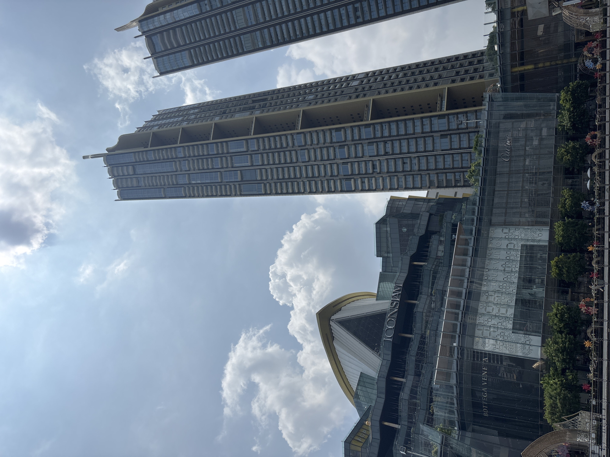 The image shows a vertical view of Bangkok, Thailand, featuring the ICONSIAM shopping mall and The Residences at Mandarin Oriental, Bangkok. ICONSIAM is recognizable by its modern architectural design and the presence of luxury brand signage like Bottega Veneta and Louis Vuitton. The two tall, modern buildings are The Residences at Mandarin Oriental, Bangkok. The sky is partly cloudy, with white clouds scattered across a light blue background. A decorative garland and several trees are visible along the edge of the image.