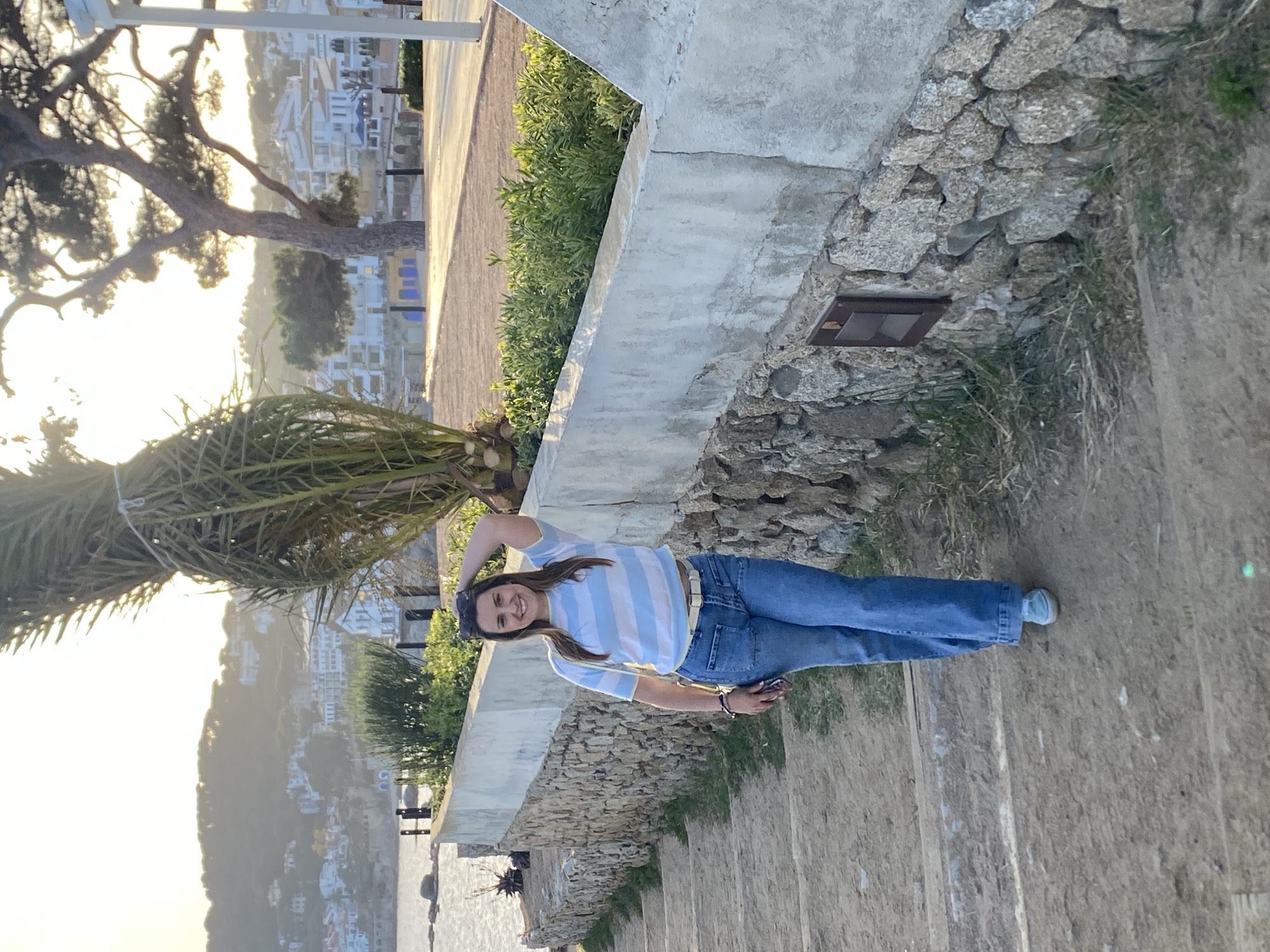 A medium shot depicts a young woman standing on an outdoor staircase, angled towards the viewer’s right. She’s wearing a blue and white striped shirt, jeans, and sneakers. She is smiling and has one hand on her head. The background suggests a seaside location in Palafrugell, Spain, evidenced by buildings on a hillside and a glimpse of the sea. A palm tree is visible on the left. The staircase is stone with vegetation alongside, and a concrete barrier separates the walkway from the scene below.