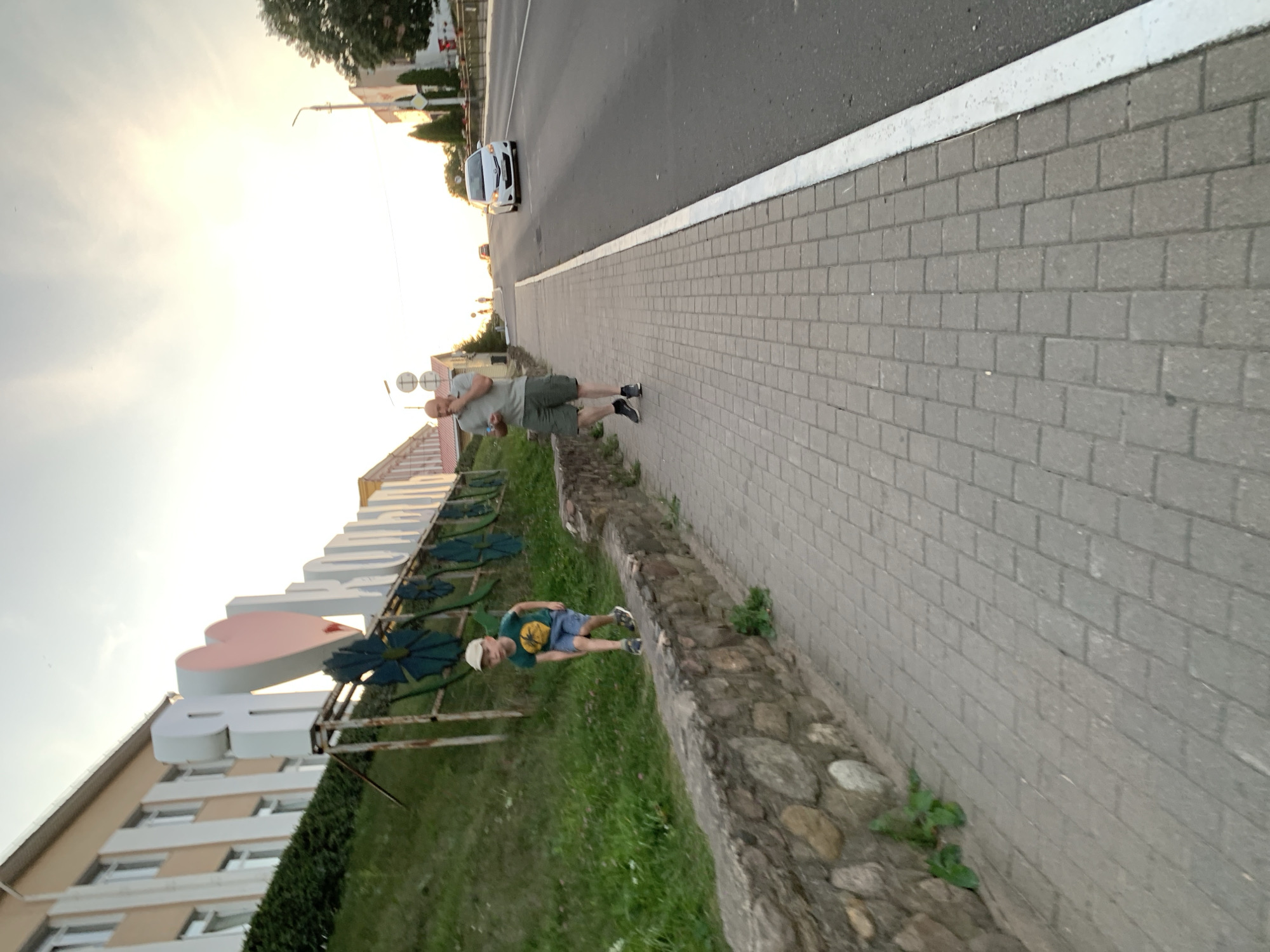 In Kapyĺ, Belarus, a child in a green t-shirt and blue shorts walks along a stone wall bordering a paved sidewalk. Behind the child, large white letters form a sign, with one letter shaped like a pink heart. A man in a grey t-shirt and shorts walks on the sidewalk further down, facing away from the camera and appearing to be smoking. The sidewalk runs alongside a road where a white car is driving. Buildings and trees line the street in the background, with a light-colored sky indicating late afternoon or early evening.
