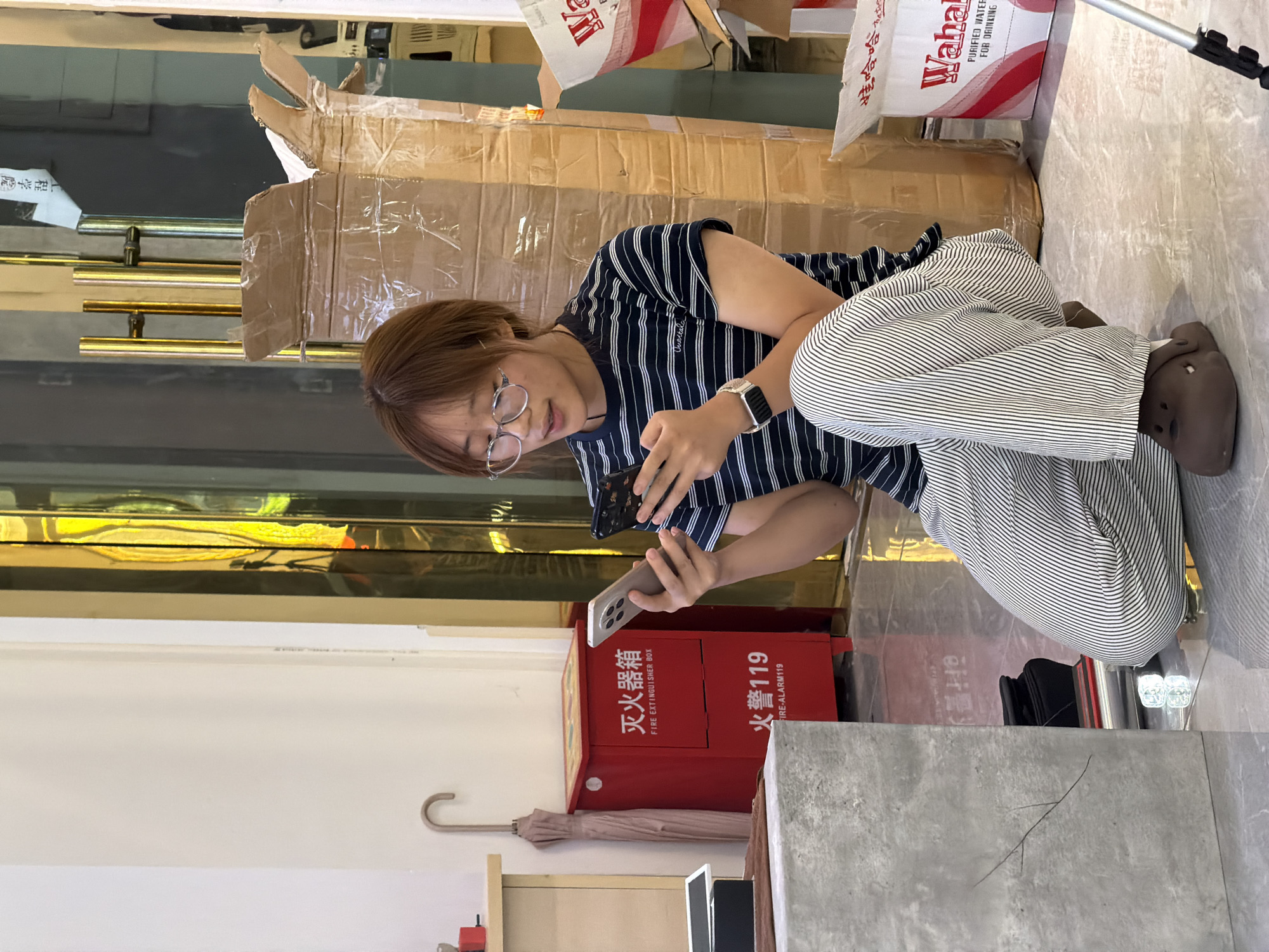 The image depicts a young woman kneeling on a tiled floor, looking at two mobile phones. She is wearing glasses, a striped t-shirt, and striped pants. In the background, there is a red fire extinguisher box with Chinese and English text, an umbrella, cardboard boxes, and what appears to be store displays. The scene is indoors, possibly in a commercial establishment, given the presence of the fire extinguisher box and packaging materials. The lighting suggests it is daytime. The image does not provide specific clues about its location in Jiangjin, China, beyond being an indoor urban environment.