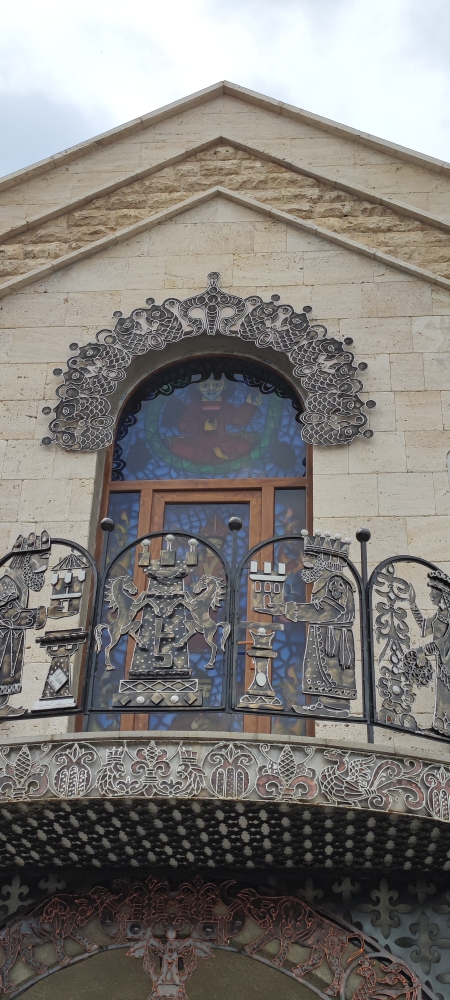 The image shows the facade of a building in Voskevaz, Armenia. The building is constructed of light-colored stone blocks. The facade features an arched window with stained glass depicting a seated figure. Above the window is ornate metalwork, resembling stylized fish or scales. Below the window, a decorative metal balcony railing displays figures and motifs, including chess pieces, horses, and human figures in elaborate attire. The balcony has intricate patterns, and below it, further metalwork with animal motifs, such as bulls, is visible. The overall aesthetic is elaborate and decorative, suggesting a building of cultural or historical significance.