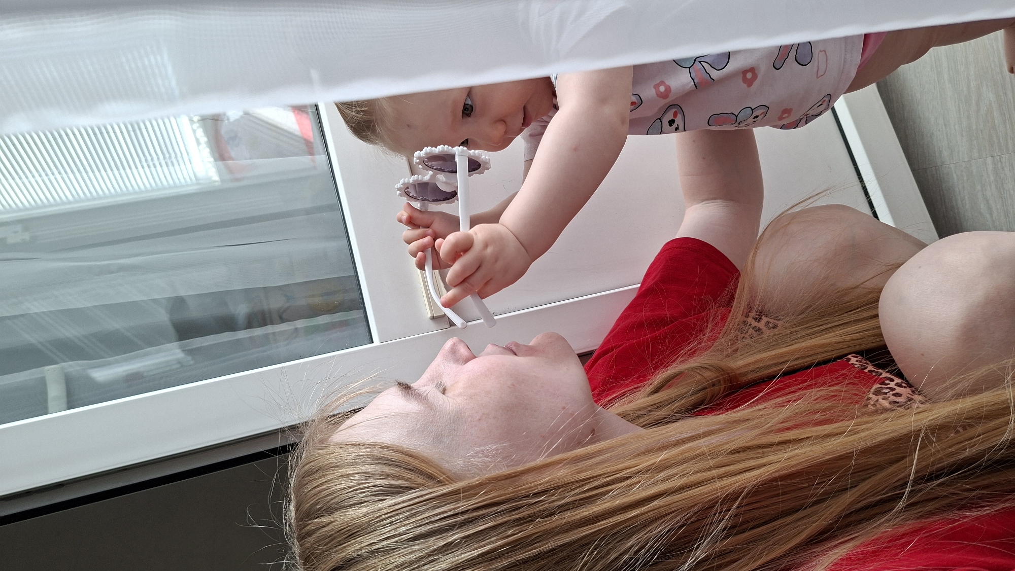 A low-angle, medium shot shows a blonde-haired woman lying on her back, with her head tilted slightly upwards, in Chaikovsky, Russia. A baby is reaching down from above, attempting to place a pair of sunglasses on the woman's face. The baby is wearing a pink and white patterned shirt. The woman is wearing a red shirt. The background includes a window with blinds and a translucent white curtain. The floor appears to be wood-toned.