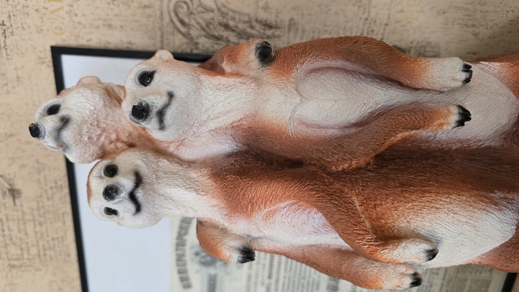 A close-up, eye-level shot shows a decorative figurine of three meerkats standing together, facing slightly to the left. The meerkats are a reddish-brown and cream color, with black noses and eyes. Their fur texture is clearly visible. The largest meerkat is in the front, with two smaller meerkats stacked behind it. The figurine is positioned in front of a wall with a textured wallpaper featuring a subtle pattern of script and decorative elements. A framed picture with a black border is visible to the left, partially obscuring the background wallpaper. Underneath the framed picture, a section of what appears to be an old newspaper or document with visible text and graphics is present. The lighting is even, highlighting the details of the figurine and its surroundings. The scene is indoors, with no indication of the specific location within the city of Верхнеказымский, Russia, other than the general context of the image source.