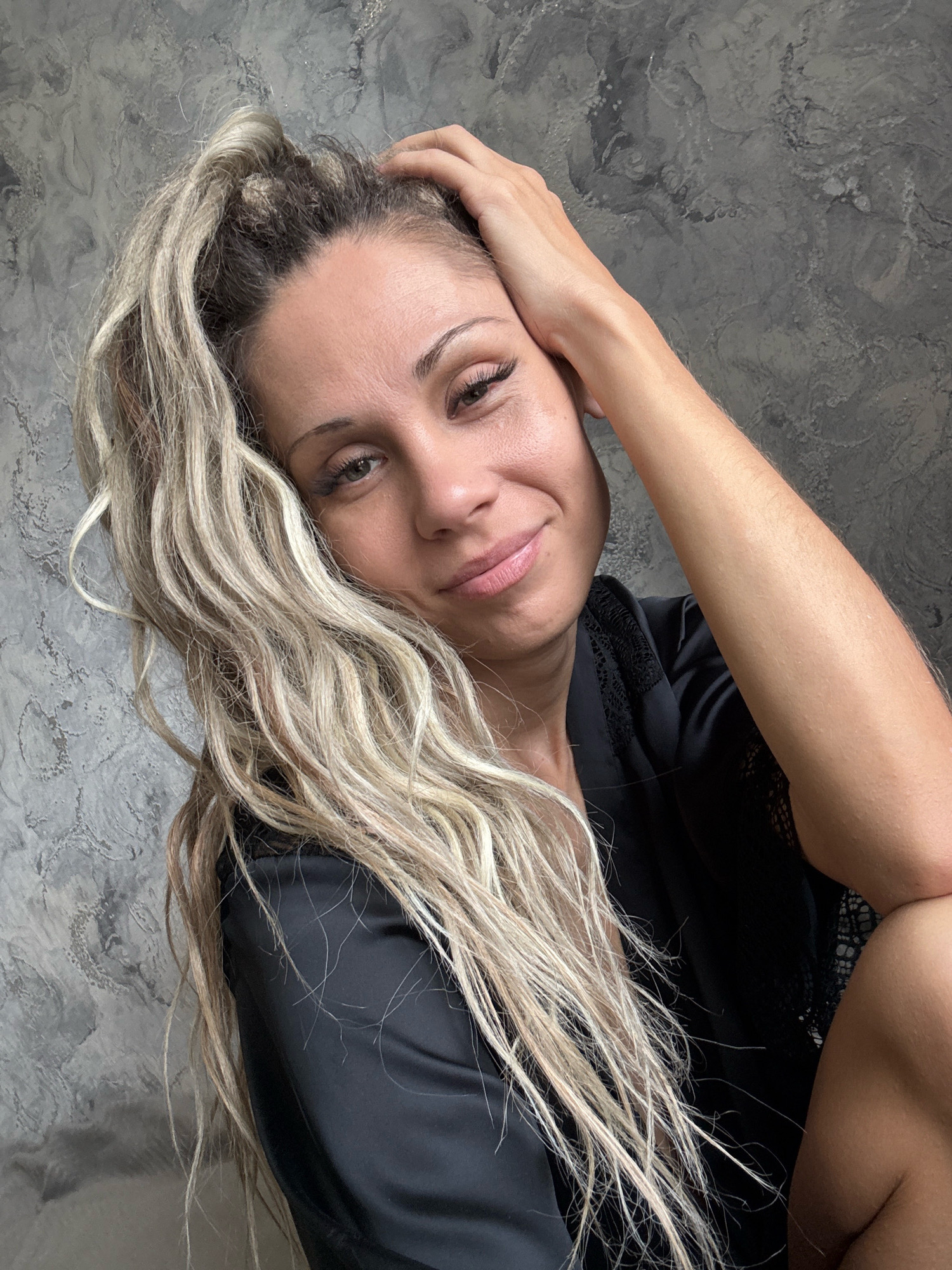 A woman with blonde hair pulled into a ponytail is leaning her head on her hand. She is wearing a black robe. The background is a textured gray wall. The image was taken in Rusanovo, Russia.