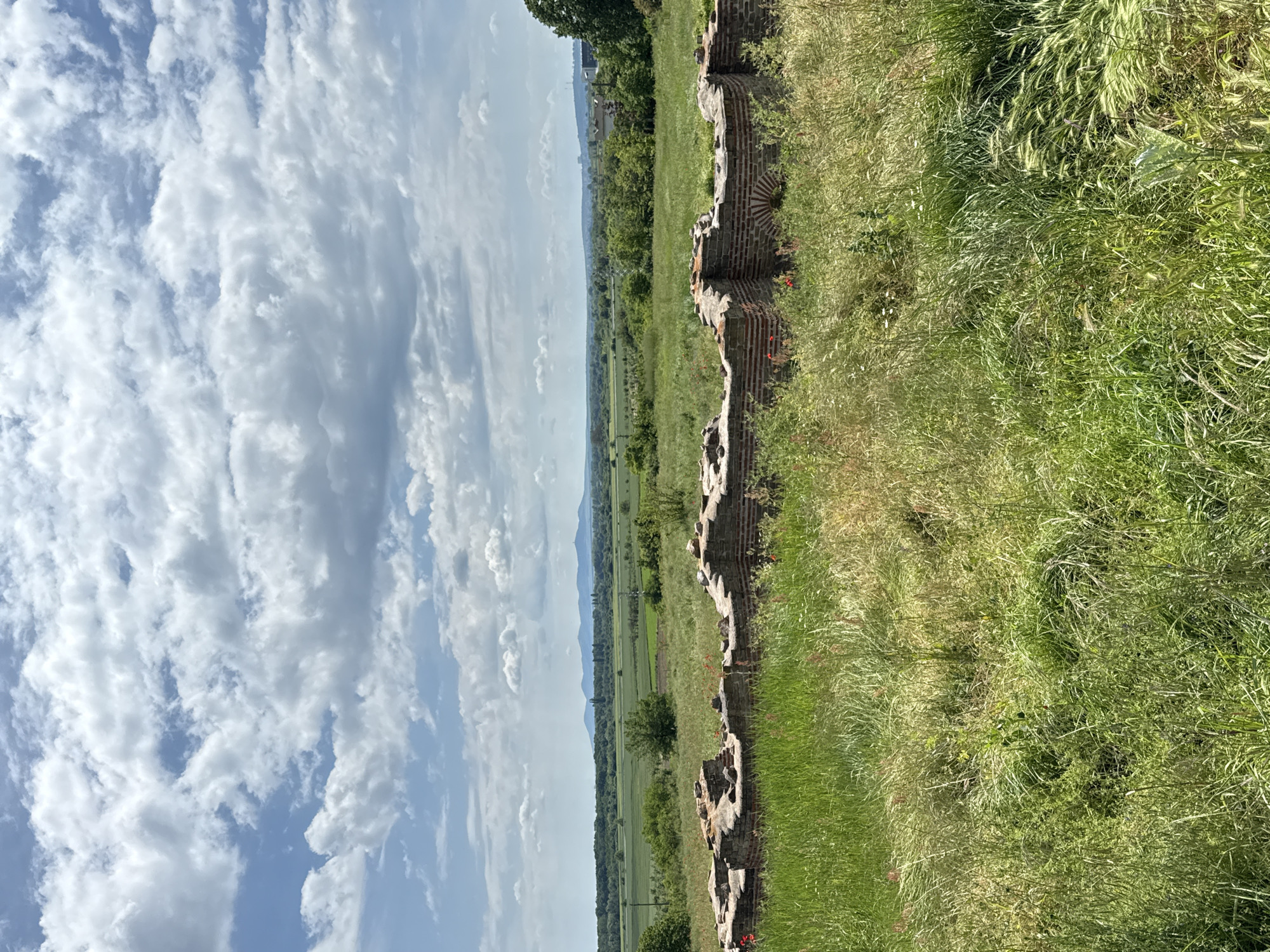 The image depicts a partially ruined brick structure, possibly a wall or part of a building, amidst a grassy field under a blue sky with scattered clouds. In the distance, a flat plain stretches to the horizon, punctuated by trees and possibly a road or pathway. There are no people visible. Based on the location mentioned, the scene could be a historical or archaeological site within the city of Kabile, Bulgaria. The bricks appear weathered, indicating age and exposure to the elements. The grassy vegetation suggests a natural environment that is not heavily maintained.