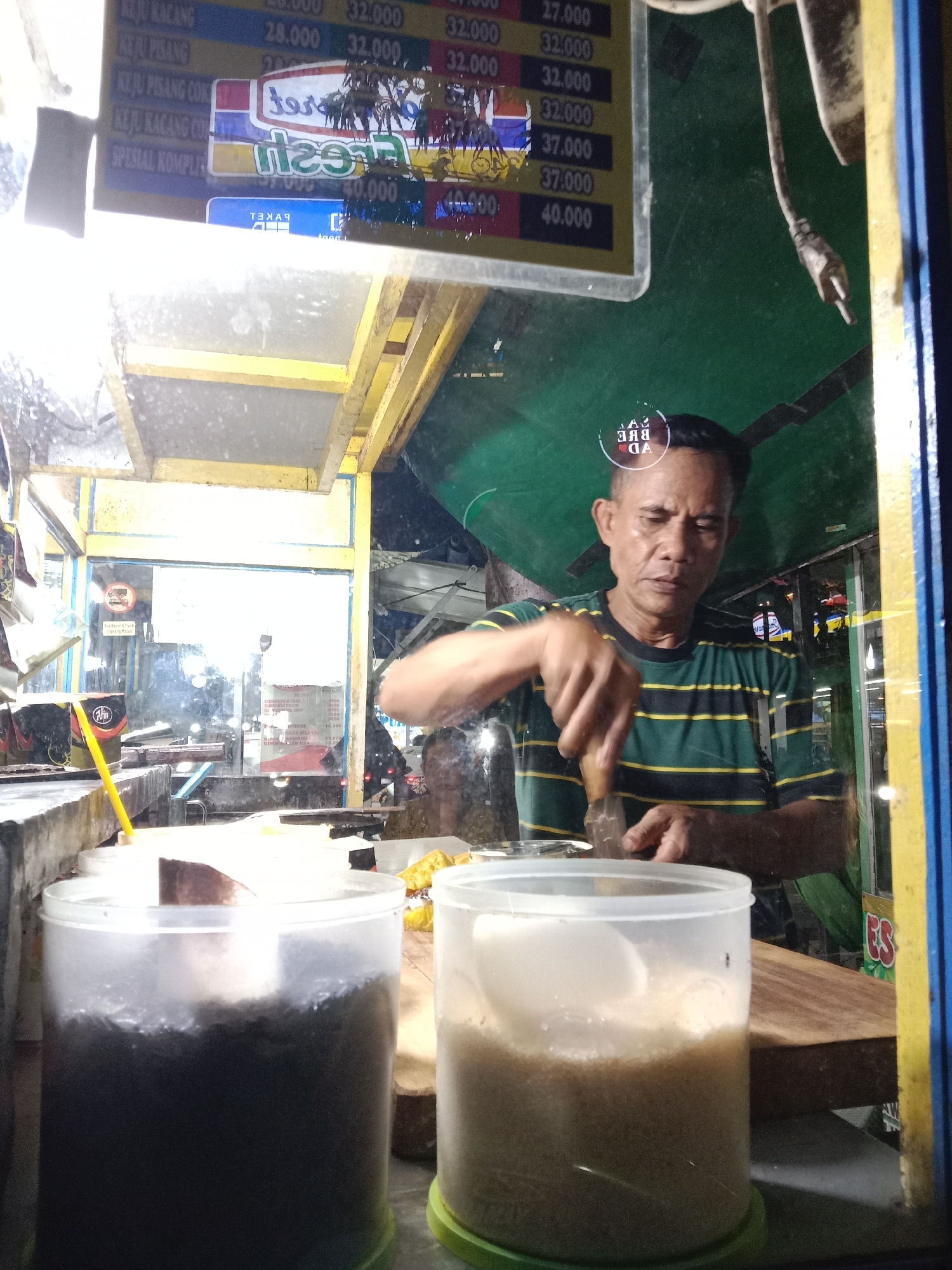 This nocturnal image captures a male vendor diligently preparing food at a street stall in Kaliwungu, Indonesia. The vendor, clad in a green and yellow striped shirt, is focused on his task, using a tool—possibly a knife or scraper—on a wooden chopping board situated behind a clear glass counter.
In the foreground, two transparent plastic containers hold ingredients or toppings: one containing a dark, granular substance and the other a lighter, sandy-textured material, both with scoops inside. The stall, characterized by its yellow frame and clear panels, reflects the surrounding environment.
Visible reflections on the glass include a menu board displaying various items and their prices in Indonesian Rupiah. Text such as "KEJU KACANG" (Cheese Peanut) and "KEJU PISANG" (Cheese Banana) are listed with prices ranging from 28,000 to 40,000. Other reflected text includes "fresh", "Alin", "TEMA", "GA", and a reversed "ЗАЯ ЯВА" on the vendor's forehead. A faint 'no trucks' sign is also discernible. The artificial lighting suggests it is night, and the overall scene depicts a typical, busy street food operation. The vendor's expression is one of concentration, suggesting dedication to his craft.