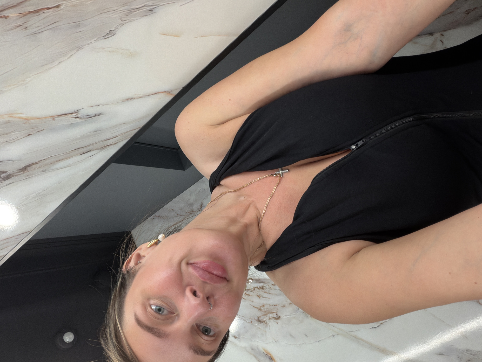 A woman with fair skin and light brown hair is taking a selfie in a bathroom with marble walls. She is wearing a black sleeveless top with a zipper and a gold necklace with a cross pendant. She has a nose ring and pearl earrings. She is smiling and looking at the camera. The background is a marble wall with gray and white veins, and a black frame around a mirror.