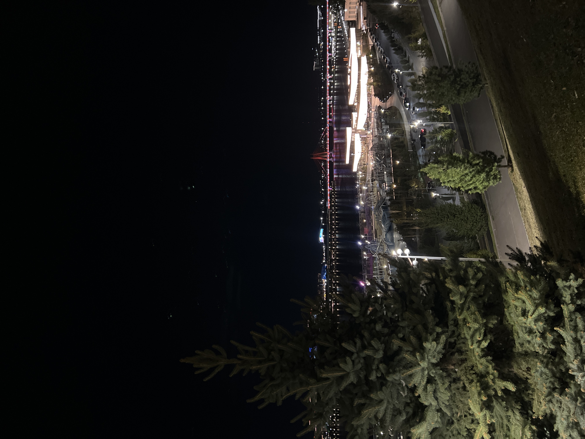 The image depicts a night scene in Kazan, Russia, likely along the Kazanka River. In the foreground, the branches of a fir tree are visible. Beyond the tree, the illuminated cityscape stretches along the riverbank. A bridge with red and white lights spans the river. Bright lights shine off of nearby buildings. The dark expanse of the Kazanka River occupies the left side of the image. On the right side, a road with parked cars runs parallel to the river. The image is oriented vertically, with the sky above.