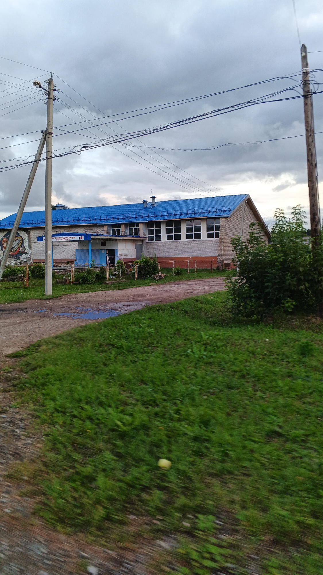 The image captures a building with a blue metal roof in the village of Podgornoye, Russia. The building appears to be a cultural center or similar public facility, indicated by signage that reads "ПОДГОРНЕНСКИЙ СЕЛЬСКИЙ ДОМ КУЛЬТУРЫ" which translates to "Podgornoye Rural House of Culture". A mural depicting a figure in what appears to be a historical or ceremonial costume is visible on the left side of the building. The structure is constructed with light-colored brick. In front of the building, there is a dirt road with puddles of water, and to the right, a dense green bush. Utility poles with electrical wires are present on the left and right sides of the frame, with a lamppost attached to the pole on the left. The sky is overcast with gray clouds. The foreground shows a blurred grassy area with a small yellow object, possibly an apple, on the ground.