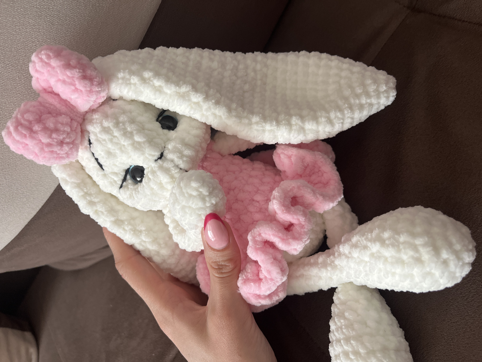 A person is holding a plush, hand-crocheted white bunny rabbit with pink accents. The bunny has long white ears, black button eyes, and a stitched black mouth. It has a pink bow on its head and a pink ruffled belly. The person's hand is visible, with a fingernail painted pink with a red tip. The background is a brown upholstered surface. It's impossible to confirm the location of the city of Pernik, Bulgaria, as there's no identifiable information in the image to support that claim.
