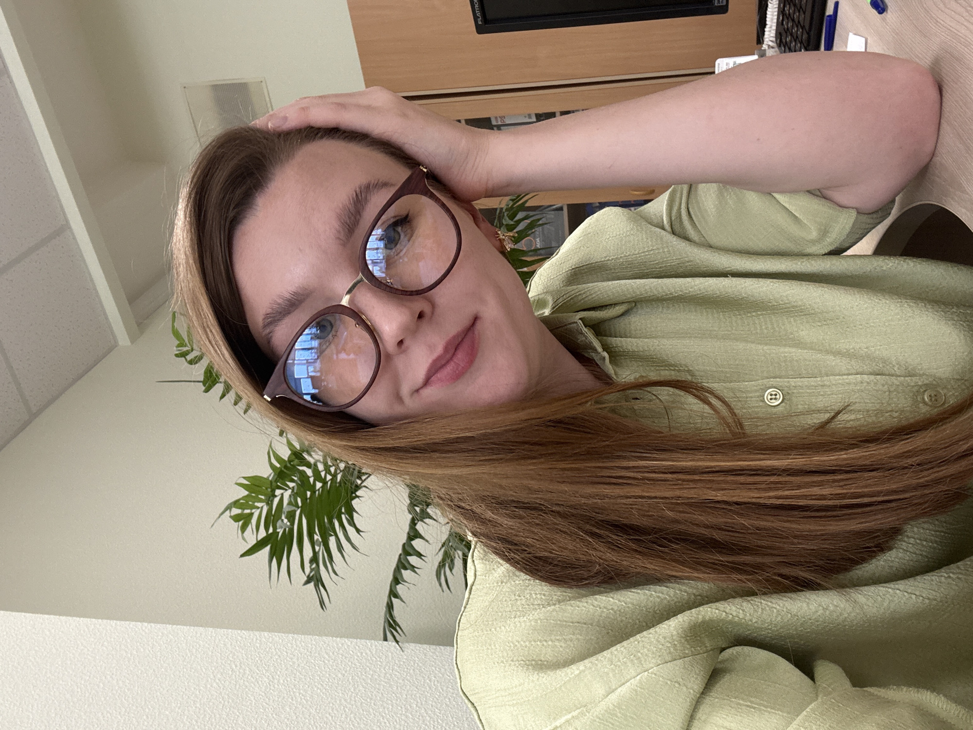 A woman with long reddish-brown hair and glasses poses for a selfie in an indoor setting. She wears a light green textured shirt with visible buttons. Her left hand is raised and rests on her head. A green leafy plant is visible behind her, along with parts of a wooden desk or shelf unit. A whiteboard or plain wall is in the background. There is nothing in the image to indicate the location is Izhevsk, Russia.