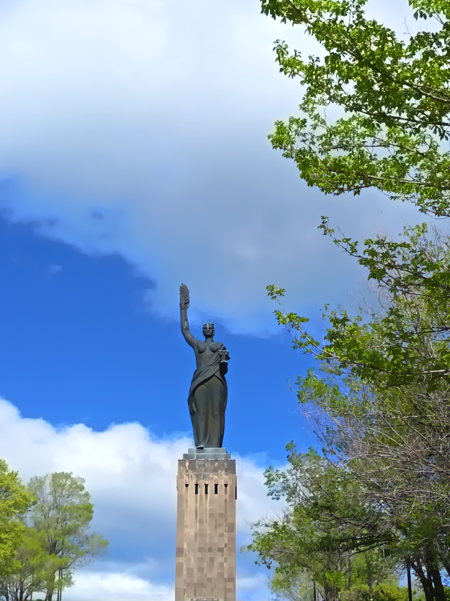 The image shows a statue of a woman holding a sheaf of wheat or a similar plant in her raised right hand. She is dressed in a long robe and standing on a tall, rectangular pedestal with vertical grooves. The statue and pedestal are located outdoors under a blue sky with scattered white clouds. Trees with green foliage are visible on both sides of the statue, framing it. The specific location within the city of Unknown, Unknown is not discernible from the image.
