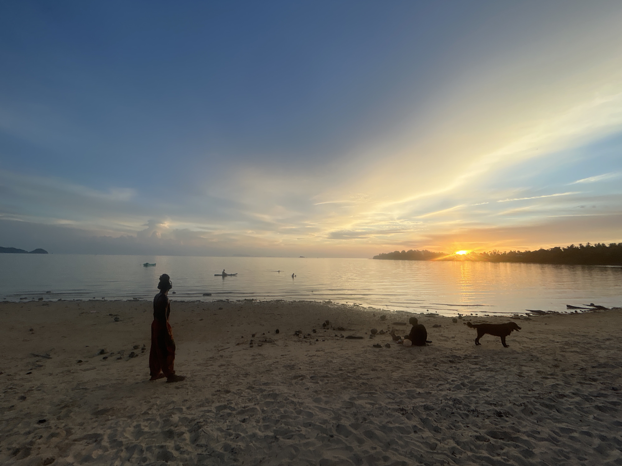 The image captures a serene beach scene in Ko Pha-ngan District, Thailand, during sunset. The sky is painted with hues of orange, yellow, and blue, with the sun setting over the calm ocean horizon. The foreground features a sandy beach with several individuals and a dog.
On the left side of the beach, a person stands silhouetted against the dimming light, wearing what appears to be loose-fitting clothing in earthy tones. Further down the beach, two figures are seated in conversation or resting, also silhouetted. A dark-colored dog is seen standing and looking towards the water.
In the shallow water near the shore, several small boats or kayaks are visible. One person is seated in a kayak, while another appears to be in the water. The water itself is calm, reflecting the colors of the sky. The distant shoreline is lined with lush green vegetation, suggesting a tropical environment. The overall atmosphere is peaceful and tranquil, typical of a beach at dusk.