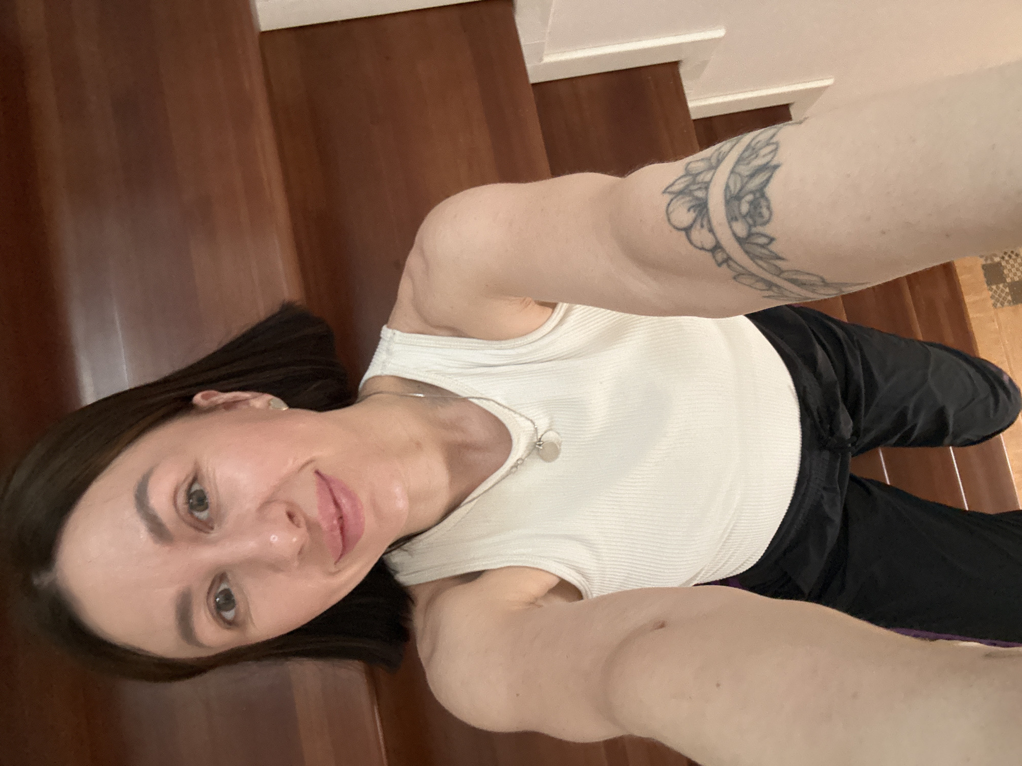 A woman is posing for a selfie while lying on a wooden floor. She is wearing a white tank top, black pants, and a silver necklace. She has a floral tattoo on her left arm. The location is a room with a wooden floor and white trim. The city of Русский Вожой, Russia, is not discernible from the image.