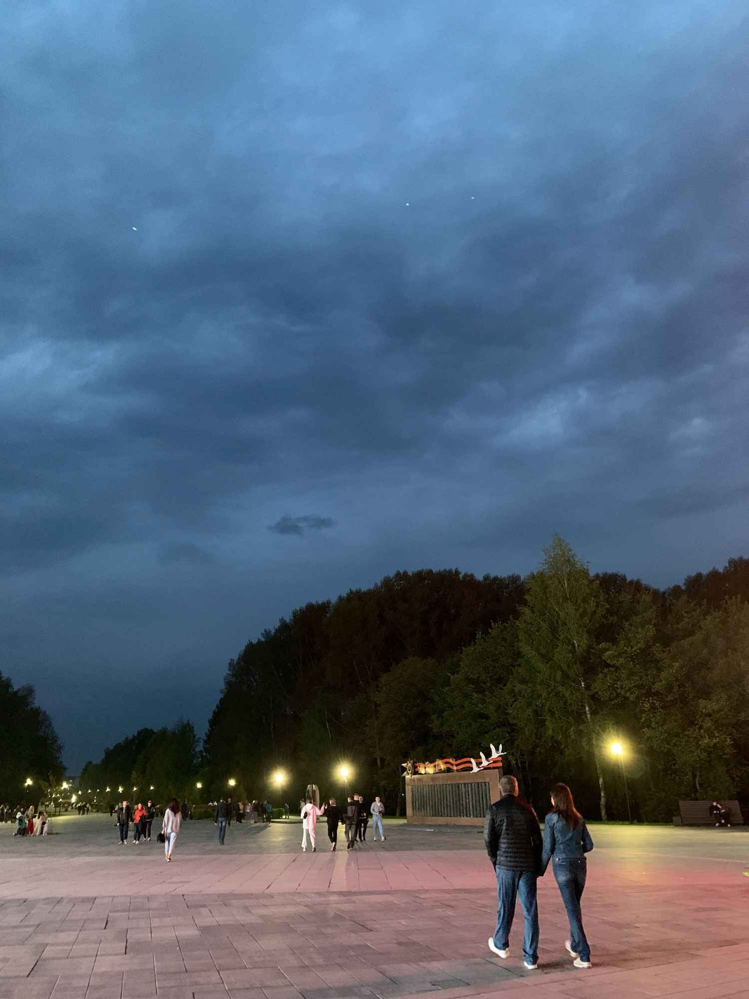 The image depicts an evening scene in Prokopyevsk, Russia, likely at Victory Park. The sky is dark with heavy, grey clouds, with two bright, small objects, possibly stars or distant lights, visible. A line of trees with dark foliage forms a backdrop along the edge of a wide, paved area. In the foreground, a couple walks away from the camera, holding hands. The man wears a dark jacket and blue jeans, while the woman wears a denim jacket and jeans. Other people are scattered across the paved area, some walking, some gathered in groups. Streetlights with a warm glow are visible along the walkway, illuminating the scene. In the background, a monument or structure with flags and images is partially visible, illuminated from behind. The ground is paved with large, rectangular stones, reflecting the artificial lights.