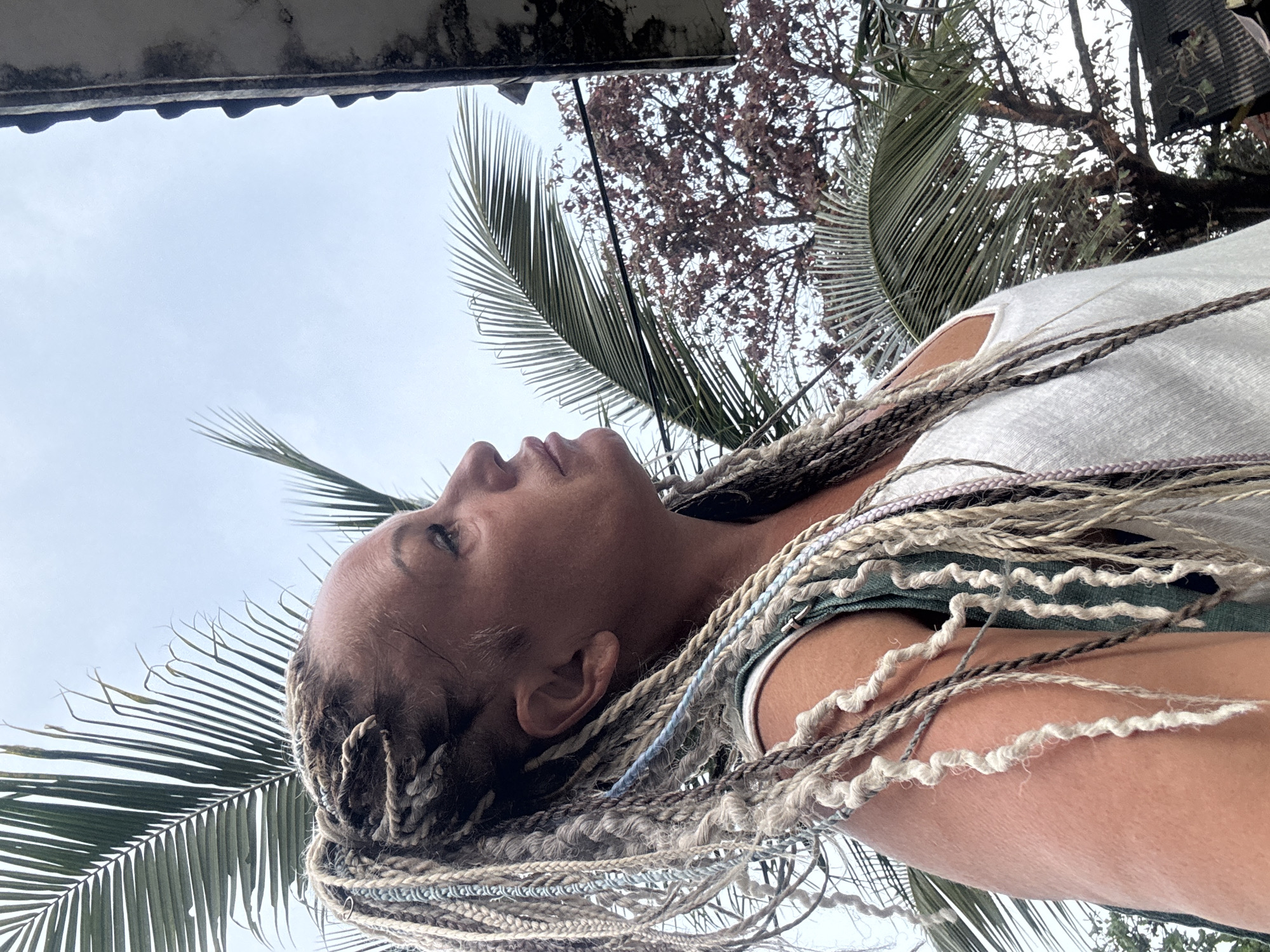 A woman with intricately braided blonde hair looks upward towards an overcast sky. The hair includes light brown, white, and even subtle shades of pale blue interwoven into the braids. She is wearing a white tank top with hints of green underclothing visible at the straps. The background includes palm fronds and branches from another type of tree. In the upper left corner, the corner of a building or structure can be seen. Without any further context, it is not possible to confirm the exact location in Kuta, Indonesia.