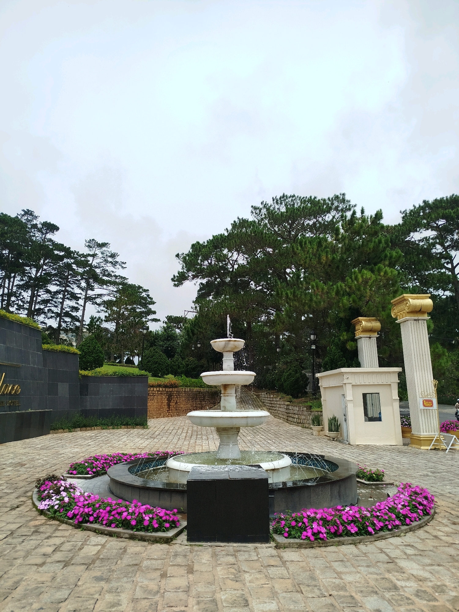 The image depicts the exterior of the Dalat Palace Hotel in Da Lat, Vietnam. In the foreground, a three-tiered white fountain with water cascading down its levels sits in a circular pool. The pool is surrounded by a raised planter filled with vibrant pink and purple flowers. The ground is paved with light-colored cobblestones.
To the left of the fountain, a dark gray, tiled wall with golden lettering, likely signage for the hotel, is visible. Behind the wall, lush green trees and manicured bushes form the hotel grounds.
To the right of the fountain, a small, cream-colored building, possibly a guardhouse or information kiosk, is situated. It has a dark-framed window. Two ornate, fluted columns with gold Corinthian capitals stand behind this building, adding a touch of classical architecture. In the background, tall pine trees and other foliage dominate the landscape, with a cloudy sky overhead. A faint figure of a person is visible in the far right background.