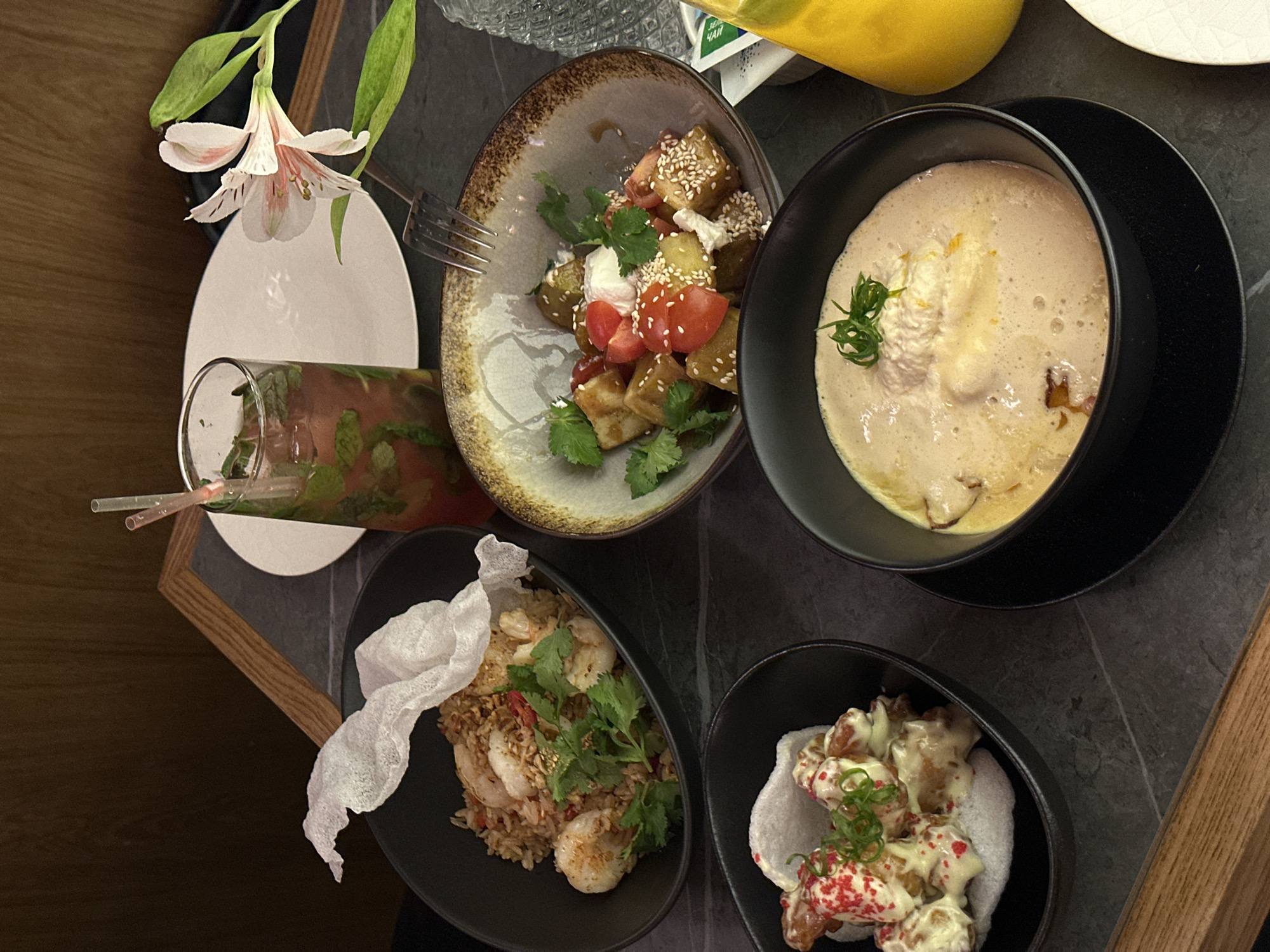 The image shows a table setting with various dishes and a drink, likely in a restaurant. The setting includes a plate with a drink (likely a cocktail) and a flower, a bowl with a salad or appetizer featuring what looks like sesame-covered fried eggplant, tomatoes and cilantro, a bowl of soup, a bowl of fried rice with shrimp, and a bowl containing what looks like fried food (likely chicken) with sauce, possibly served on a rice cracker. The table is dark, possibly made of stone or a dark material, and framed by dark and wooden edges. The food is artfully arranged and garnished. The lighting suggests an indoor environment. The description includes no reference to Rostov-on-Don.