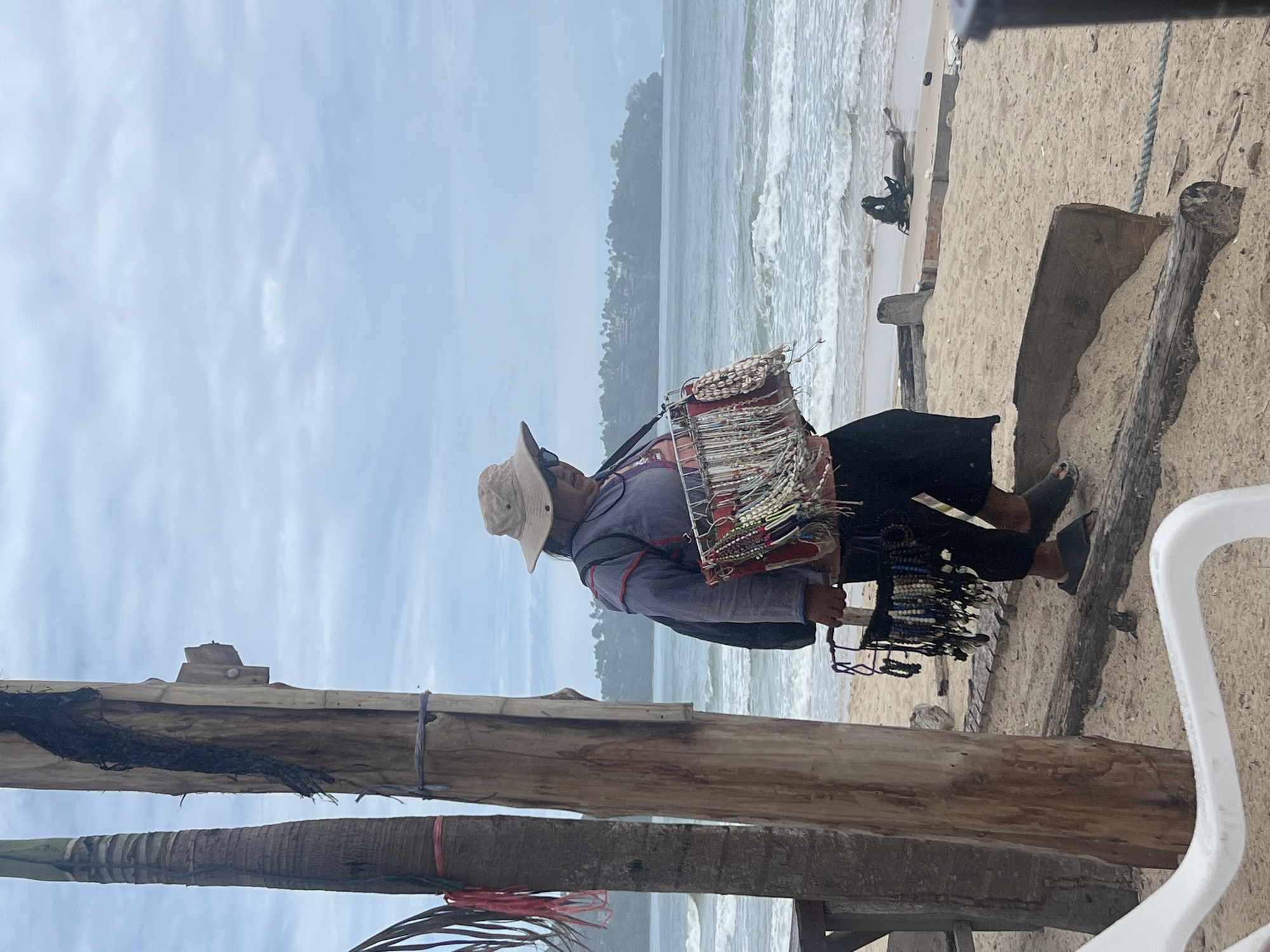A person wearing a light-colored wide-brimmed hat and sunglasses stands on a sandy beach in Choeng Thale, Thailand. They are wearing a purple shirt and black pants, and are carrying a basket filled with necklaces and bracelets. The person is walking towards the right side of the frame. In the background, the ocean is visible with waves breaking on the shore, and a line of trees and buildings can be seen in the distance. Two palm trees are to the left of the frame, and a wooden post with some twine is between them.