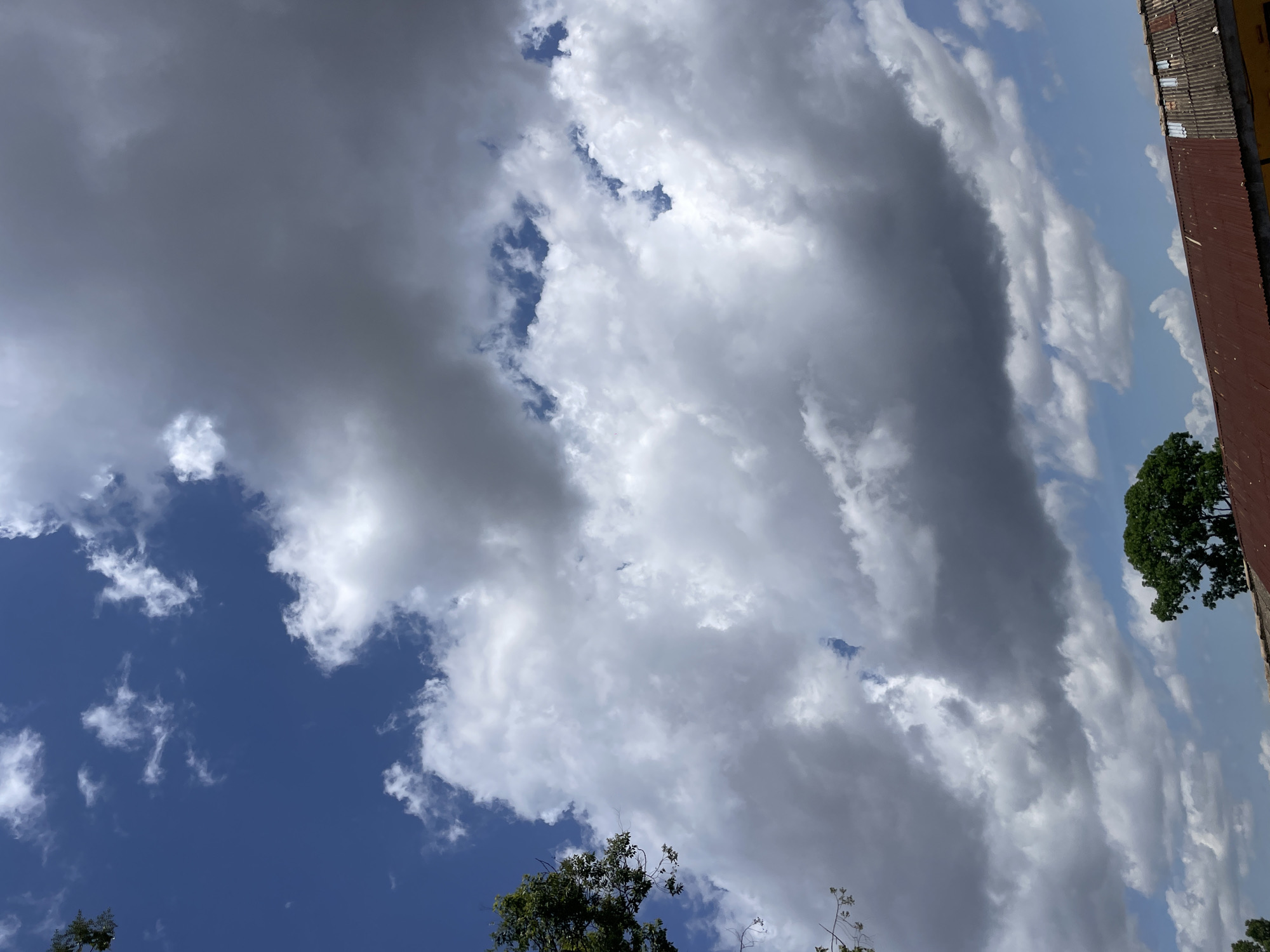 The image captures a portion of the sky over Rafin Par, Nigeria, characterized by a mix of bright blue sky and dramatic cloud formations. Dense, white cumulus clouds dominate the upper half of the frame, interspersed with patches of darker, more ominous clouds that suggest potential rain. A tree with green foliage is visible in the upper left corner, its branches extending into the frame. In the lower portion of the image, a weathered, reddish-brown corrugated metal roof of a building is visible, with glimpses of yellow paint on the wall beneath it. A large, dense green tree stands to the right of the roofline. The scene is set outdoors under natural daylight.