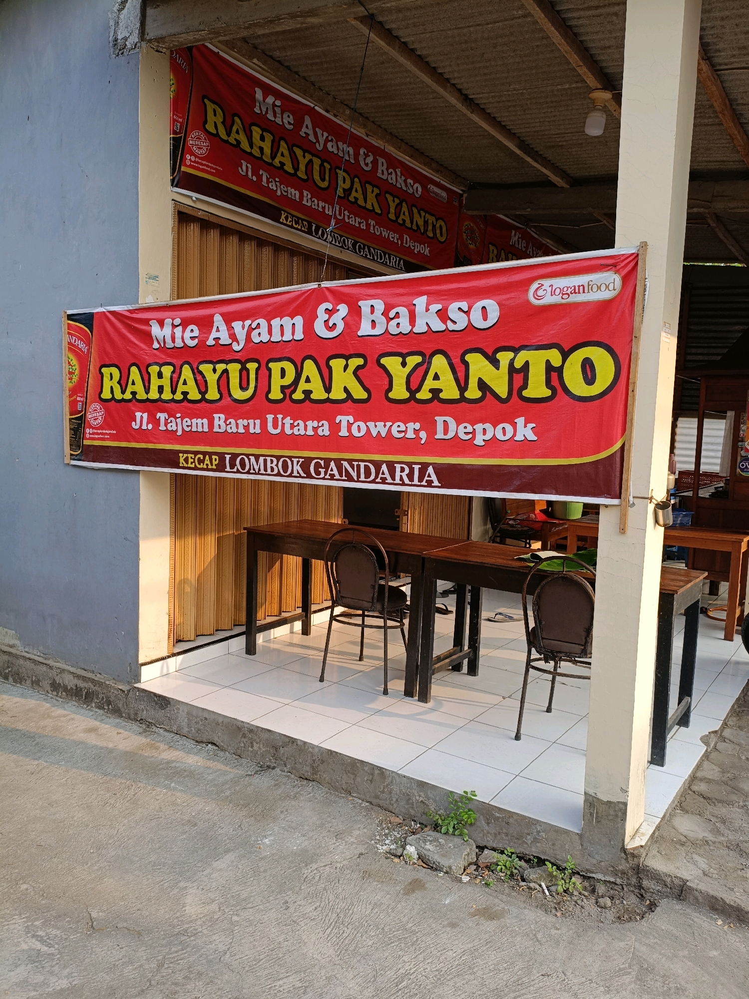 The image shows an outdoor dining area of a food establishment, likely a noodle and meatball restaurant, indicated by the prominent red banner that reads "Mie Ayam & Bakso RAHAYU PAK YANTO" with the address "Jl. Tajem Baru Utara Tower, Depok" and "KECAP LOMBOK GANDARIA". The establishment appears to be located in the city of Maguwoharjo, Indonesia, as suggested by the contextual information. The banner is hung across the front of a building with a light-colored wall on the left and a corrugated metal roof supported by wooden beams above. A cream-colored pillar stands to the right of the banner. In the foreground, there are several dark wooden tables and chairs set up on a tiled floor. The tables are empty, and the chairs are neatly placed around them, suggesting the establishment is either closed or between customers. Sunlight casts shadows across the scene, indicating it is daytime. The overall environment appears to be a casual, open-air dining space, with a slightly weathered aesthetic, as seen in the concrete pavement and the building's exterior.