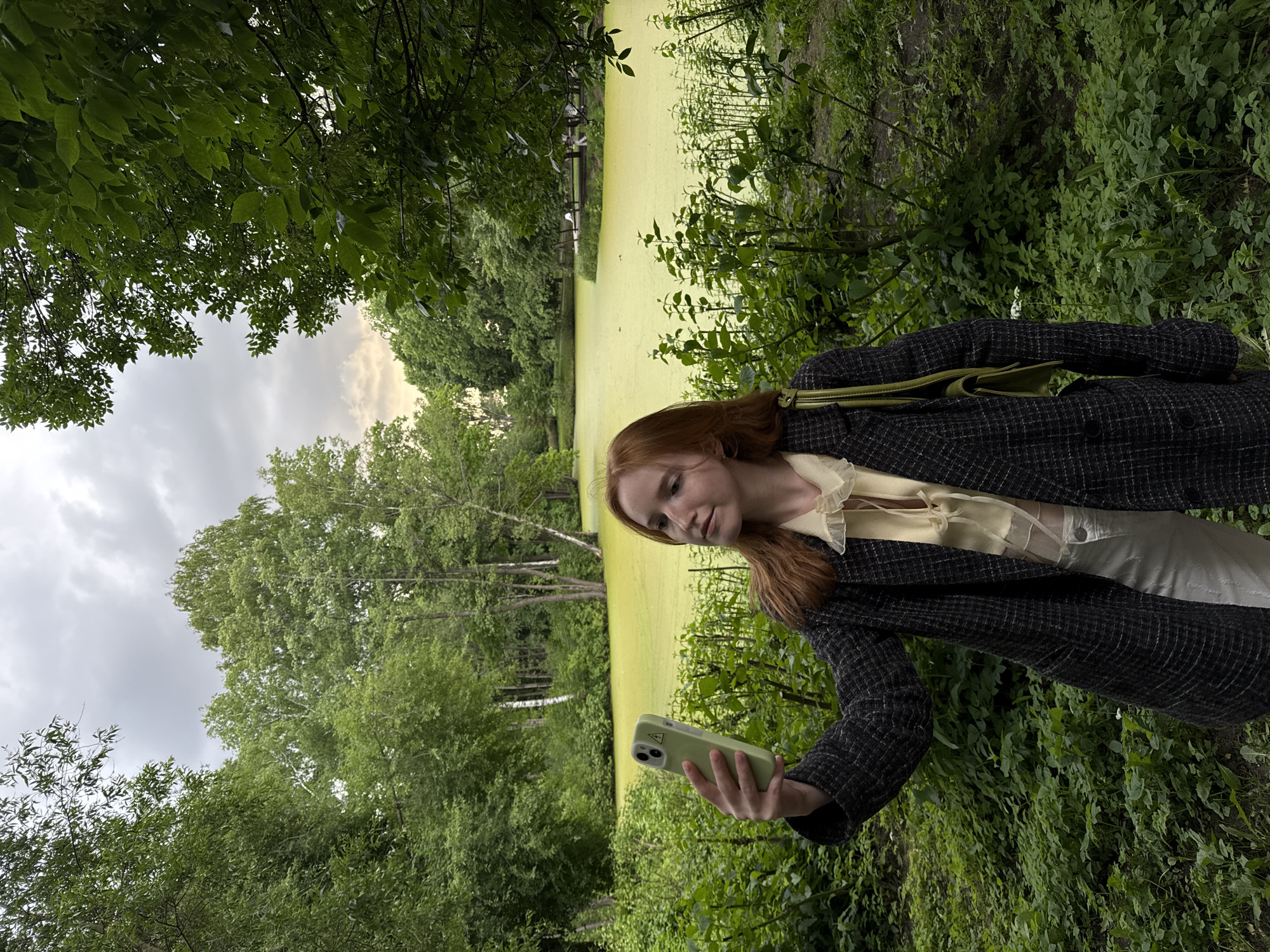 A young woman with red hair is taking a selfie with her phone in a park. She is wearing a dark, checkered jacket over a light yellow blouse and white pants. A green purse is slung over her shoulder. She is standing in front of a pond covered in bright green algae, surrounded by lush green trees and foliage. The sky is overcast with hints of yellow from the setting sun. The scene appears to be from a park in Saint Petersburg, Russia.
