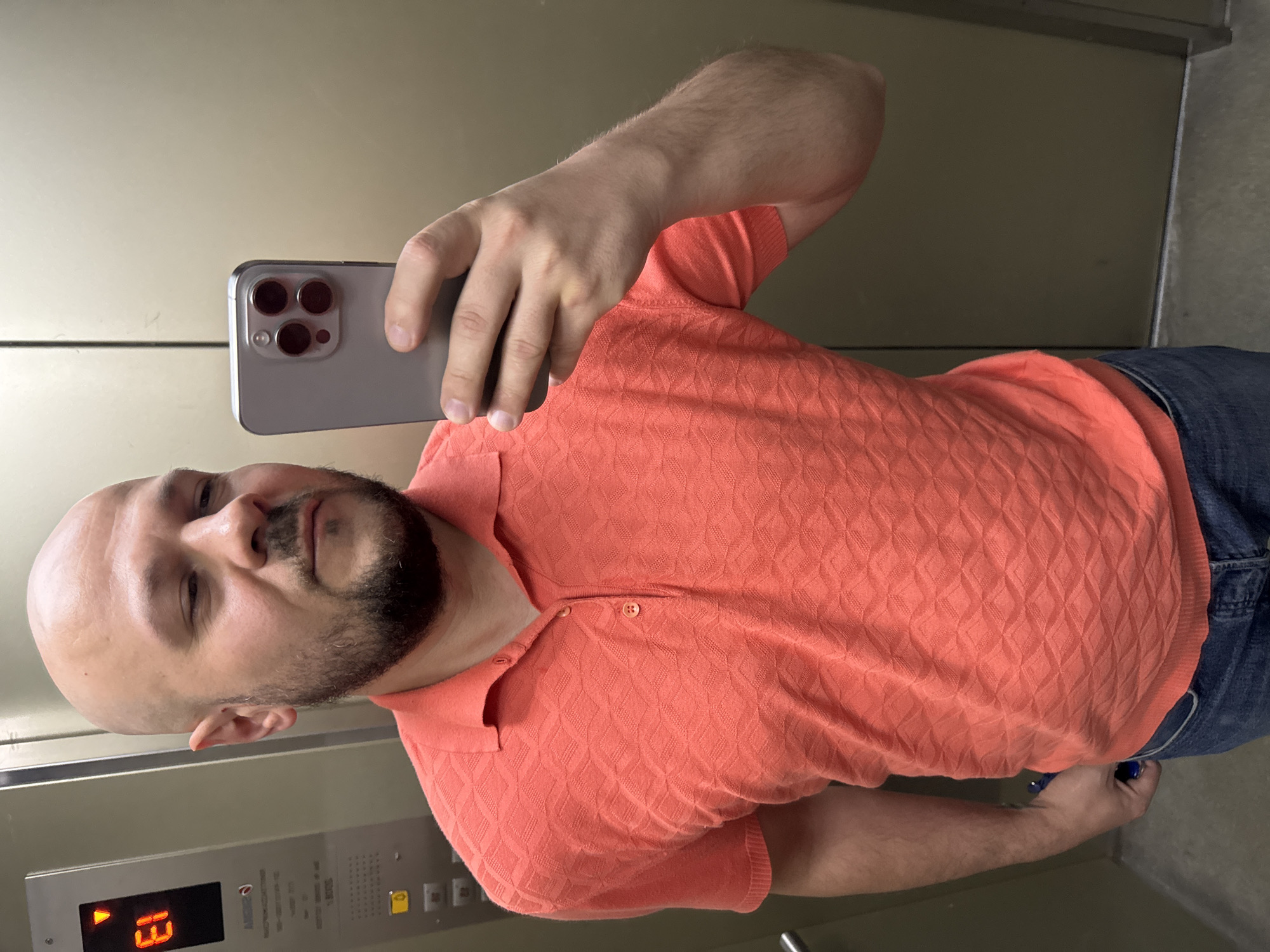 A bald, white male with a short beard and light skin takes a selfie in an elevator. He is wearing a coral-colored, short-sleeved polo shirt with a diamond-shaped pattern and blue jeans. He is holding a silver smartphone in his right hand, angled to capture his reflection. The elevator panel is visible on the left side of the image, displaying "E1" and an upward-pointing arrow. The walls of the elevator appear to be made of a beige, metallic material. The man's left hand is visible near his hip, with a glimpse of a dark object, possibly a key or fob, peeking out from his jeans pocket. The scene suggests the man is in an urban environment, possibly a residential or office building, within the city of Almaty, Kazakhstan.