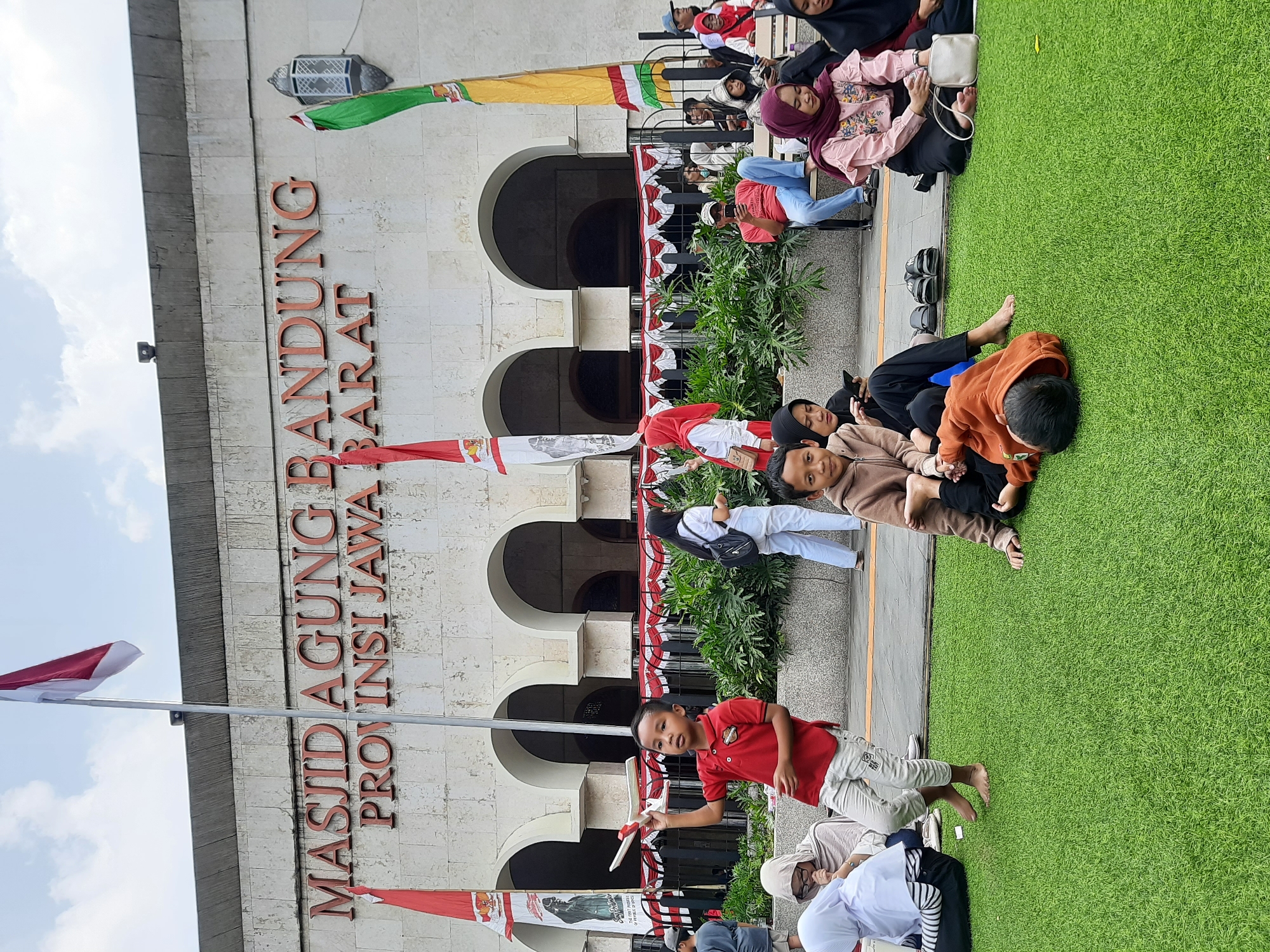 The image depicts the Masjid Agung Bandung (Grand Mosque of Bandung), a prominent religious building in Bandung City, Indonesia. The mosque's facade, constructed from stone blocks, displays the building's name in large, red, block letters: "MASJID AGUNG BANDUNG PROVINSI JAWA BARAT". Two Indonesian flags, red and white, are prominently displayed, one to the left and one to the right of the building's entrance, alongside a green and yellow flag. In front of the mosque, a group of people are gathered on a patch of artificial grass. A young boy in a red polo shirt and light-colored pants is holding a toy airplane and looking towards the camera. He is barefoot. To his right, another boy, wearing a brown hooded sweatshirt and dark pants, is lying on his back on the grass, appearing to be resting or playing. Other individuals are seated or standing around the area, some looking at their phones, while others are engaged in conversation. The scene suggests a public gathering or event taking place outside the mosque.