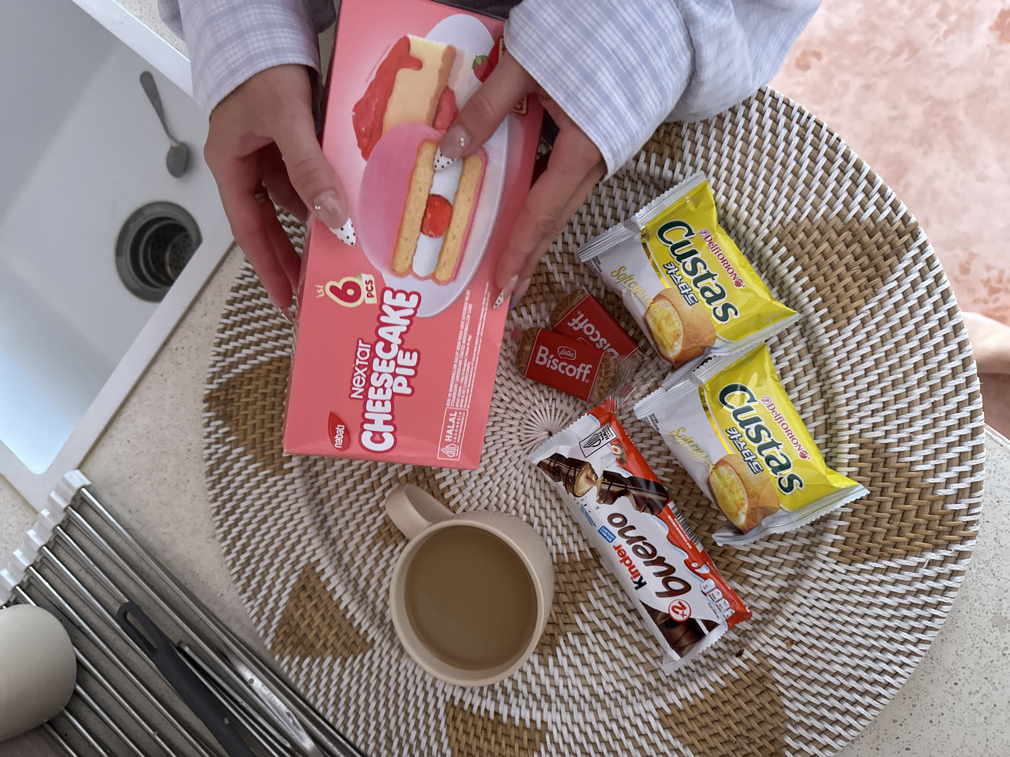 Here is a forensic description of the image:
The image shows a person holding a pink box of "Nextar Cheesecake Pie" (6 pieces) in their hands. Next to it on a woven mat are two packages of "Custas" (Delfi Orion brand), a "Lotus Biscoff" snack, and a "Kinder Bueno" bar. A mug filled with a light brown beverage is also present on the mat. The scene is set on a kitchen countertop, evident from the sink and dish rack in the background. The label on the "Nextar" product indicates "HALAL INDONESIA", which suggests the possible location in North Kuta, Indonesia.