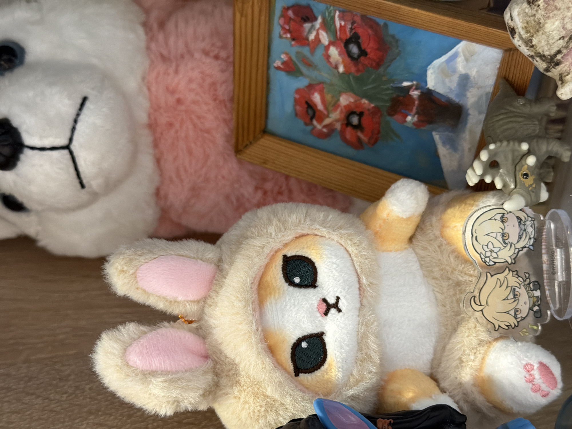 The image shows a collection of objects on a wooden surface. In the foreground, a plush bunny toy with pink ear pads and a tan hooded jacket lays on its back. Its face features black eyes and a small, pink "X" for a nose and mouth. To the left, a larger white plush bear with a pink fur section is partially visible. Above the bunny, a small framed painting of red flowers in a vase sits. To the right of the painting, a grey toy dinosaur and two anime-style character charms in a plastic case are visible. The setting is unknown but potentially someone's home. Засечное, Russia, is not explicitly apparent in this photo.