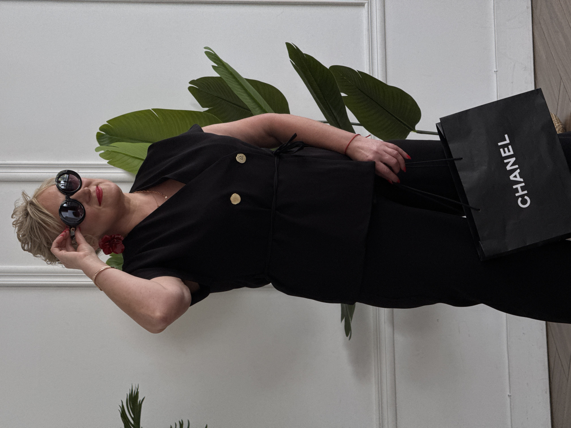 An indoor, horizontally oriented shot captures a stylish woman with blonde hair, fair skin, and red lipstick, posing against a white wall with decorative molding. She wears oversized black sunglasses, which she adjusts with her left hand. Her ensemble includes a black top or dress with a tied waist and prominent gold buttons, a gold chain necklace, a gold bracelet on her left wrist, and a thin red string bracelet on her right. A striking red floral earring adorns her right ear, and her fingernails are painted red. In her right hand, she holds a black shopping bag with "CHANEL" written vertically in white capital letters.
Behind the woman, a large potted plant with broad green leaves is visible, adding a natural element to the refined setting. The overall scene suggests a fashionable moment, possibly after a shopping excursion, within a chic and well-lit interior, located in Chaikovsky, Russia. The woman's expression, partially obscured by sunglasses, conveys a sense of confidence and style.