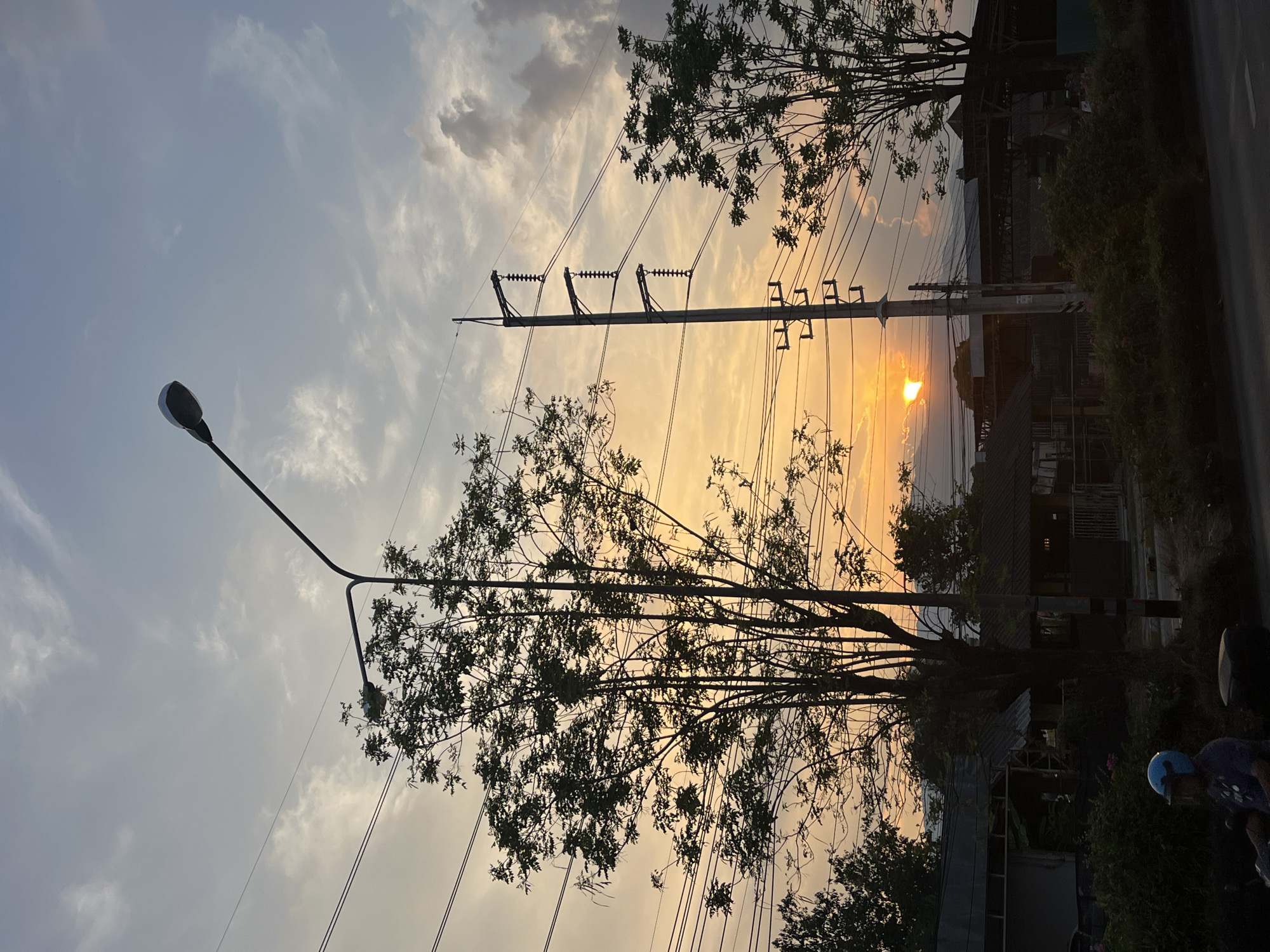 The image, taken in what appears to be a residential area in บ้านกาญจน์กนก 23, Thailand, shows a dusk or dawn scene with a sun partially obscured by buildings and trees. A utility pole with high-voltage power lines is prominently positioned in the center, accompanied by a street light on the left. Silhouetted trees frame the utility pole and partially obscure the sky. A portion of a street and buildings is visible on the right side of the image. A person wearing a blue helmet is partially visible on the bottom right. The overall impression is one of urban infrastructure intermixed with natural elements during a time of day with a distinct golden light.