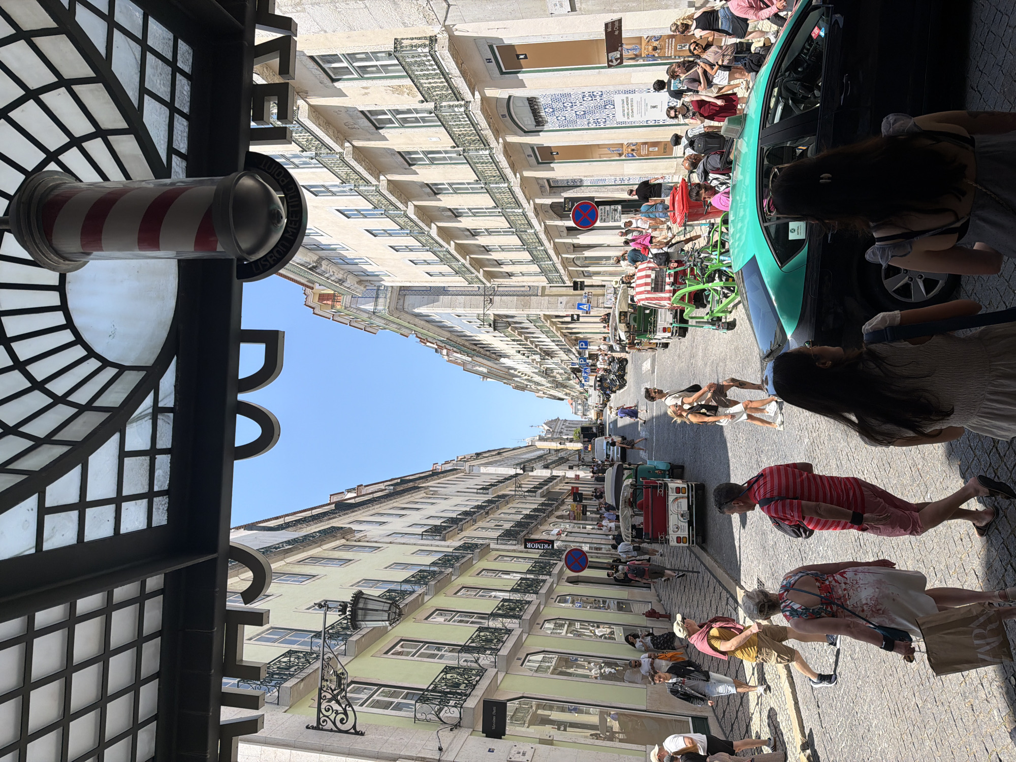 The image captures a bustling street scene in Lisbon, Portugal, likely within the Baixa district, known for its grid-like street layout and neoclassical architecture. The perspective is from beneath an awning with a black metal frame and a segmented glass ceiling, from which a classic barber pole hangs. Sunlight streams down, illuminating a cobblestone street lined with tall, narrow buildings on both sides. These buildings feature a mix of light yellow and cream-colored facades, with many windows adorned with decorative black iron balconies.
Numerous people are walking along the street, engaging in various activities. Some are tourists, evidenced by their casual attire and the presence of sightseeing vehicles like a red and white tuk-tuk or tourist cart. Others appear to be shoppers, with a few carrying shopping bags, one with a "ZARA" logo visible. A black car with a teal roof is parked on the right side of the street, and a red bin is placed near it.
The atmosphere suggests a lively commercial area on a bright, sunny day. The architectural style, street layout, and the presence of iconic Lisbon street elements like the tuk-tuks and the cobblestone streets strongly indicate this is a popular tourist area in the heart of Lisbon.